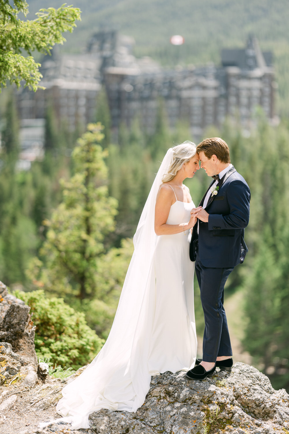 A bride and groom sharing a tender moment on a rocky outcrop with lush greenery and a large building in the background.