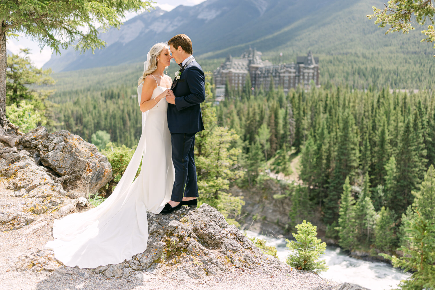 A bride and groom embracing on a rocky overlook with a majestic mountain resort and dense forest in the background.