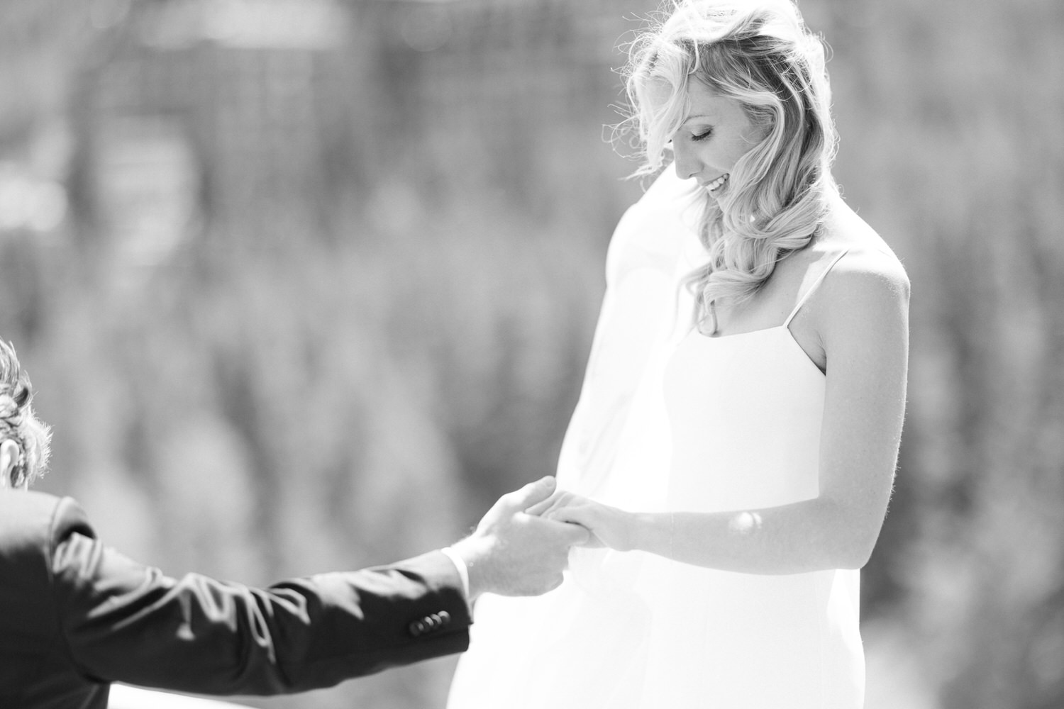 A black and white photo of a bride holding hands with her partner during a sunny wedding ceremony.