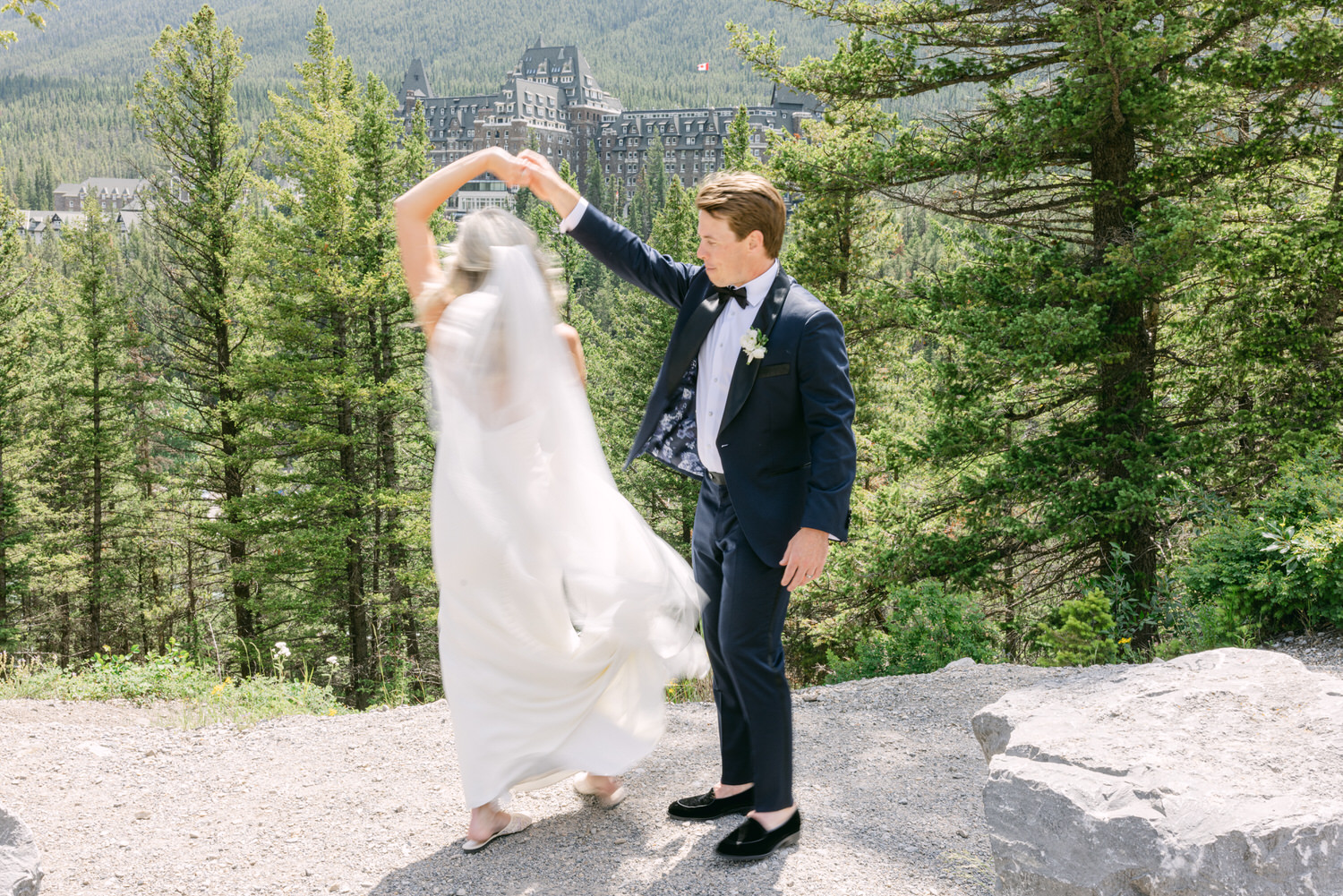 A bride and groom joyfully dance outdoors with a grand hotel in the background, surrounded by lush greenery.