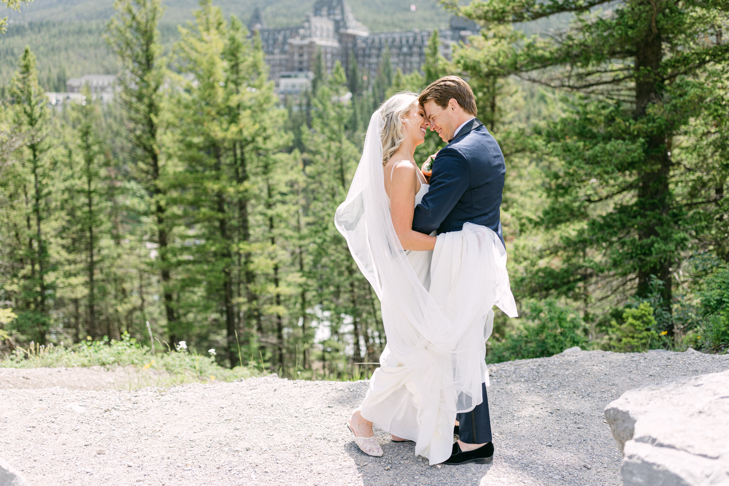 A bride in a white dress and a groom in a blue suit embracing with a scenic mountain resort backdrop.