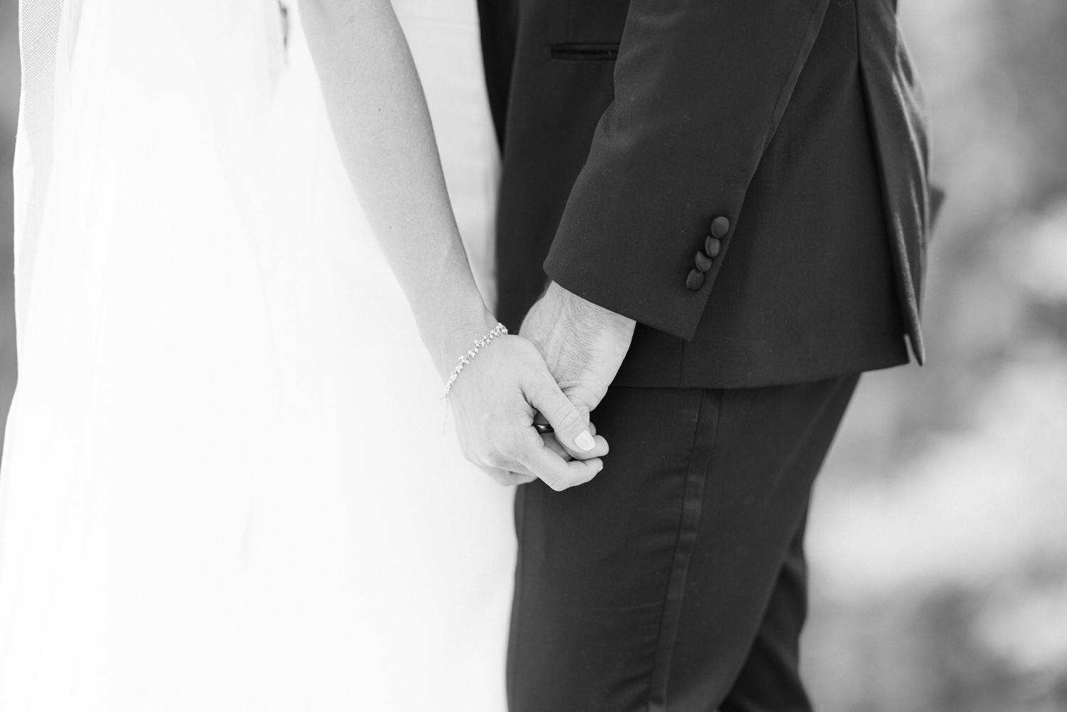 A close-up black and white photo of a newlywed couple holding hands, focusing on their joined hands with details of the wedding dress and suit.