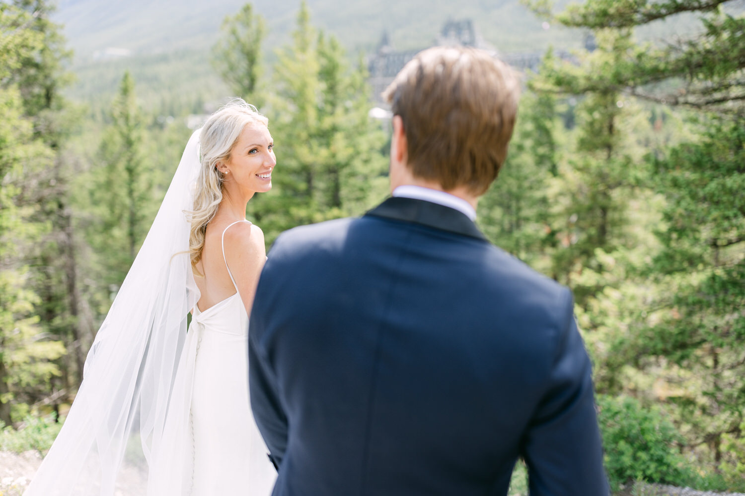 A bride in a white dress with a veil smiling at the groom in a blue suit, with lush greenery and mountains in the background.