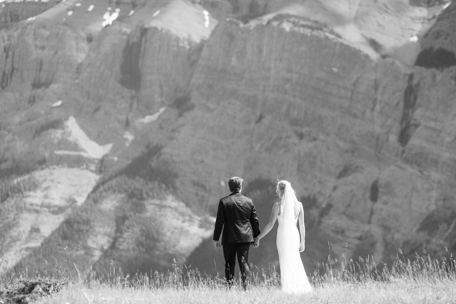 A bride and groom holding hands and walking through a grassy field with majestic mountains in the background, in black and white.