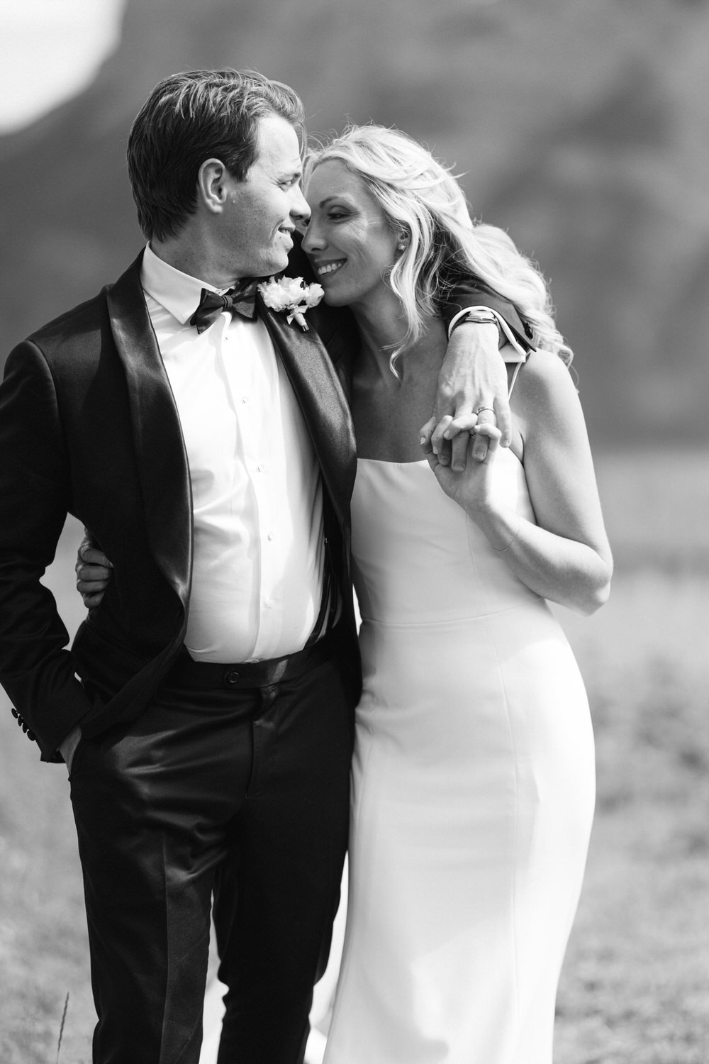 Black and white photo of a smiling bride and groom in formal attire, holding hands and looking at each other with affection.