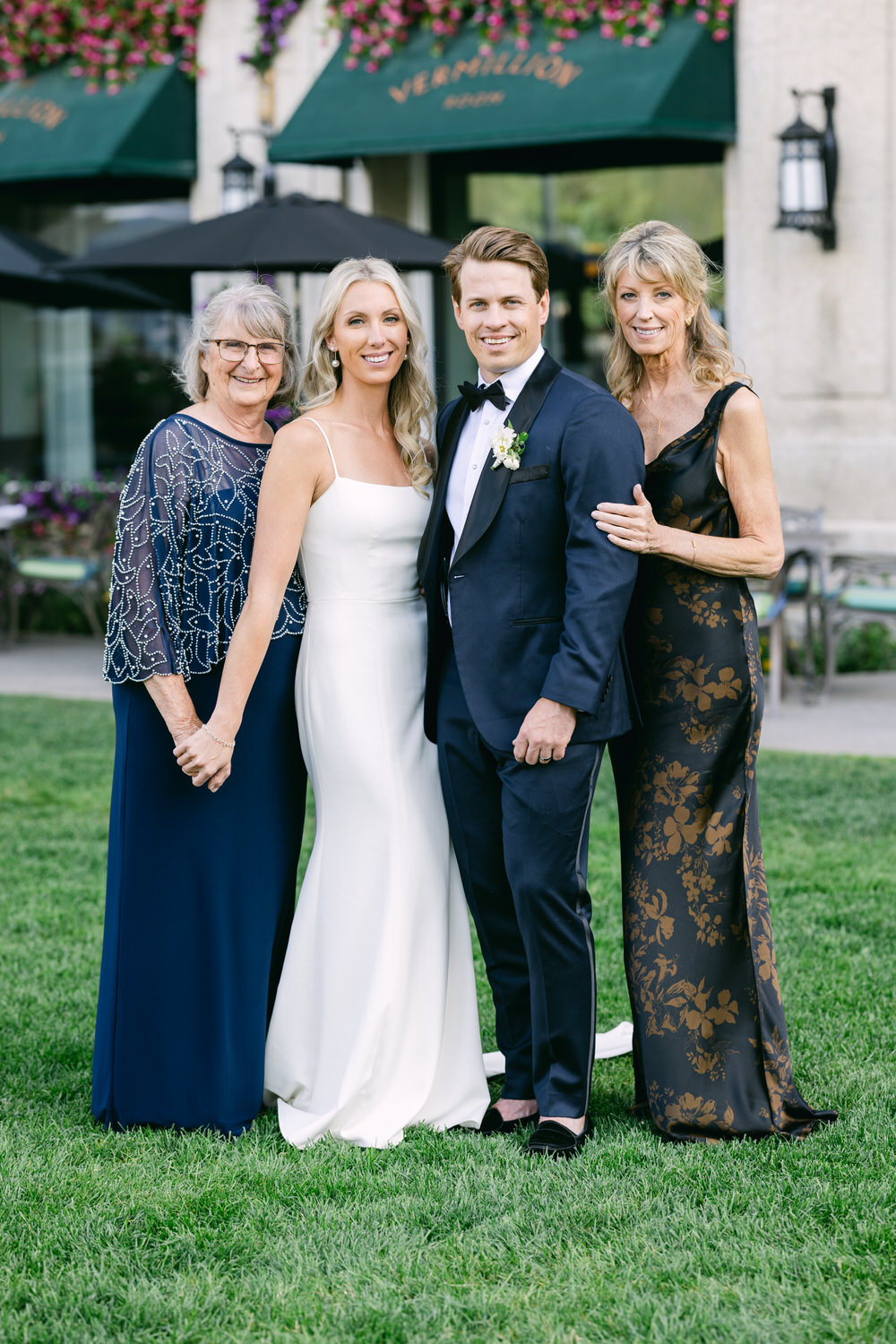 A bride and groom posing alongside two older women, all dressed in formal attire, with a restaurant facade in the background.