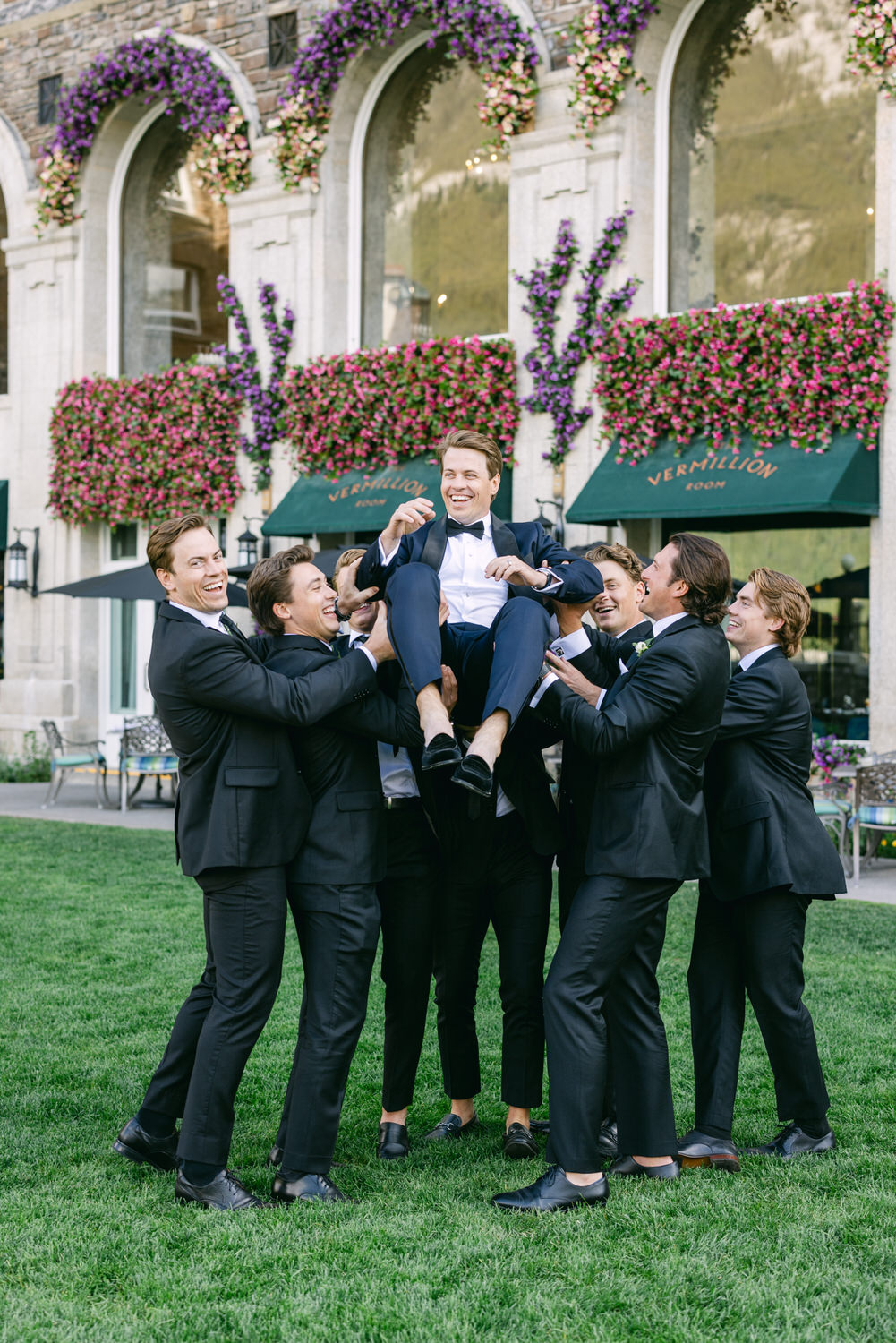 A group of groomsmen in black suits lifting the smiling groom outdoors with a flower-covered archway in the background.