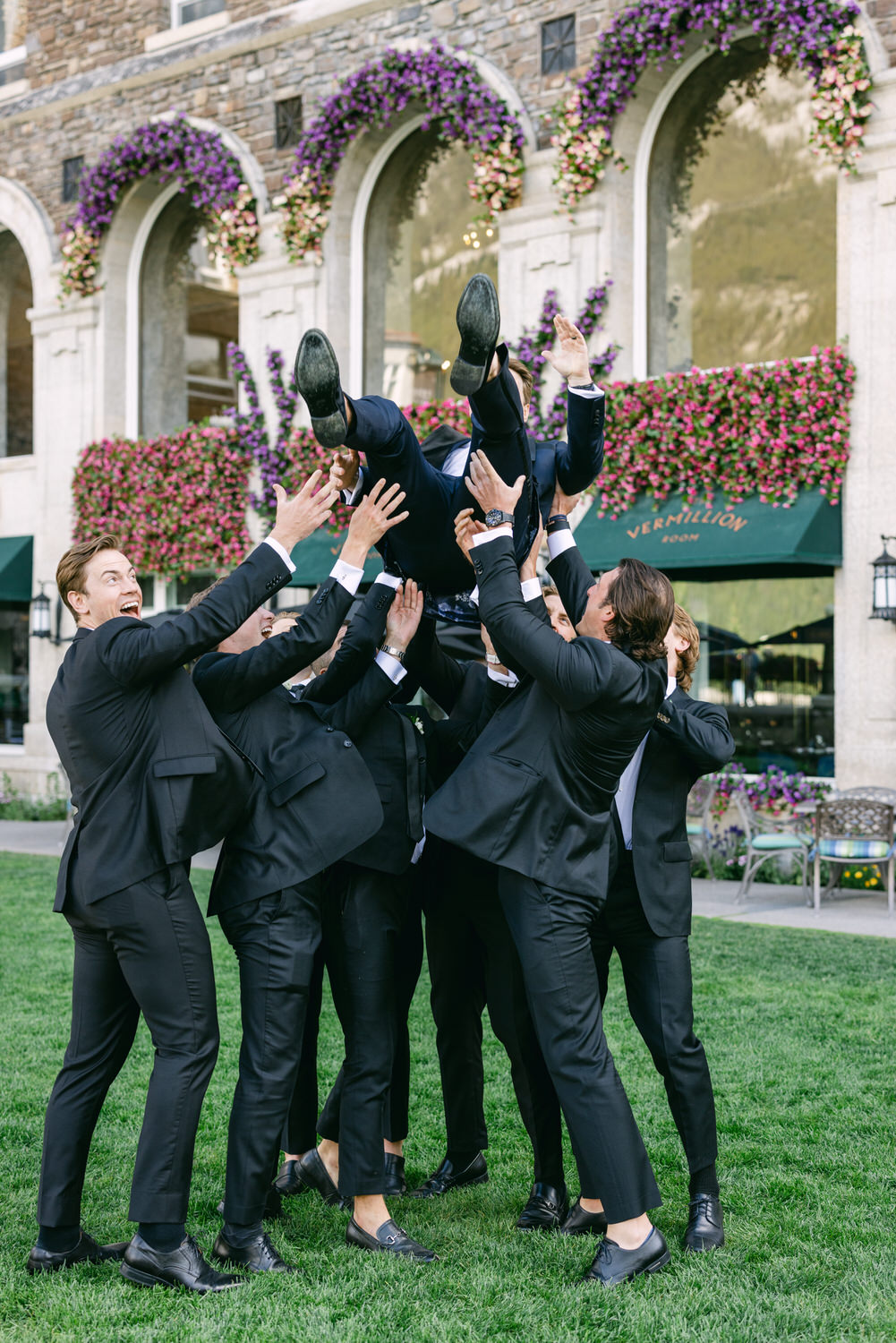 A group of groomsmen in black suits joyfully tossing the groom into the air with his feet up, in front of a building adorned with purple flowers.