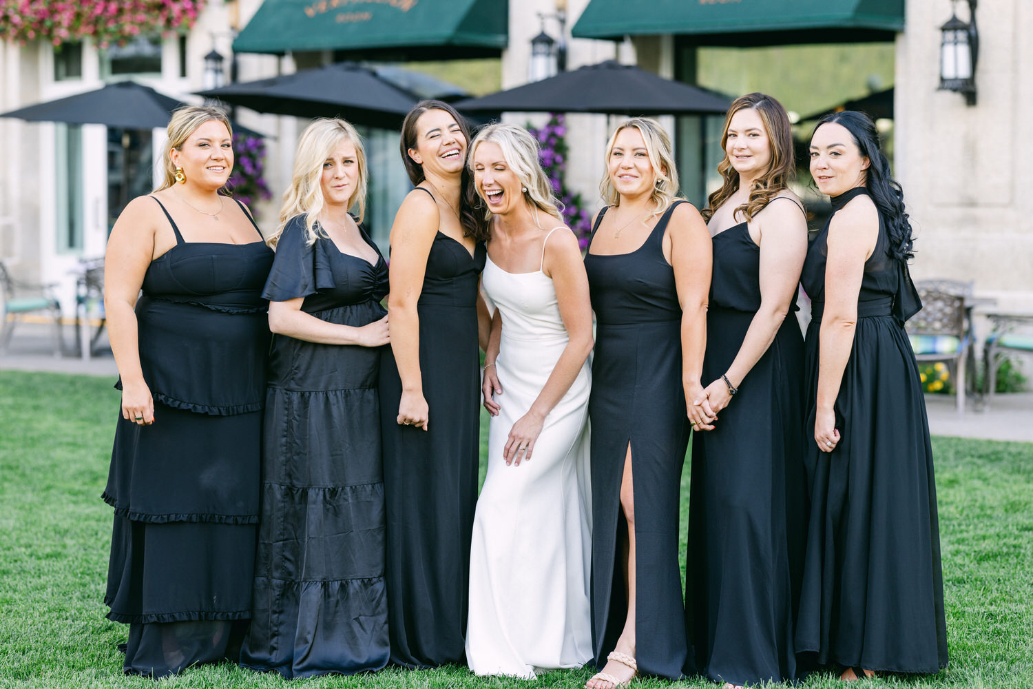 A group of women dressed in black smiling and posing together with one wearing a white dress, likely the bride, on a grassy outdoor background.