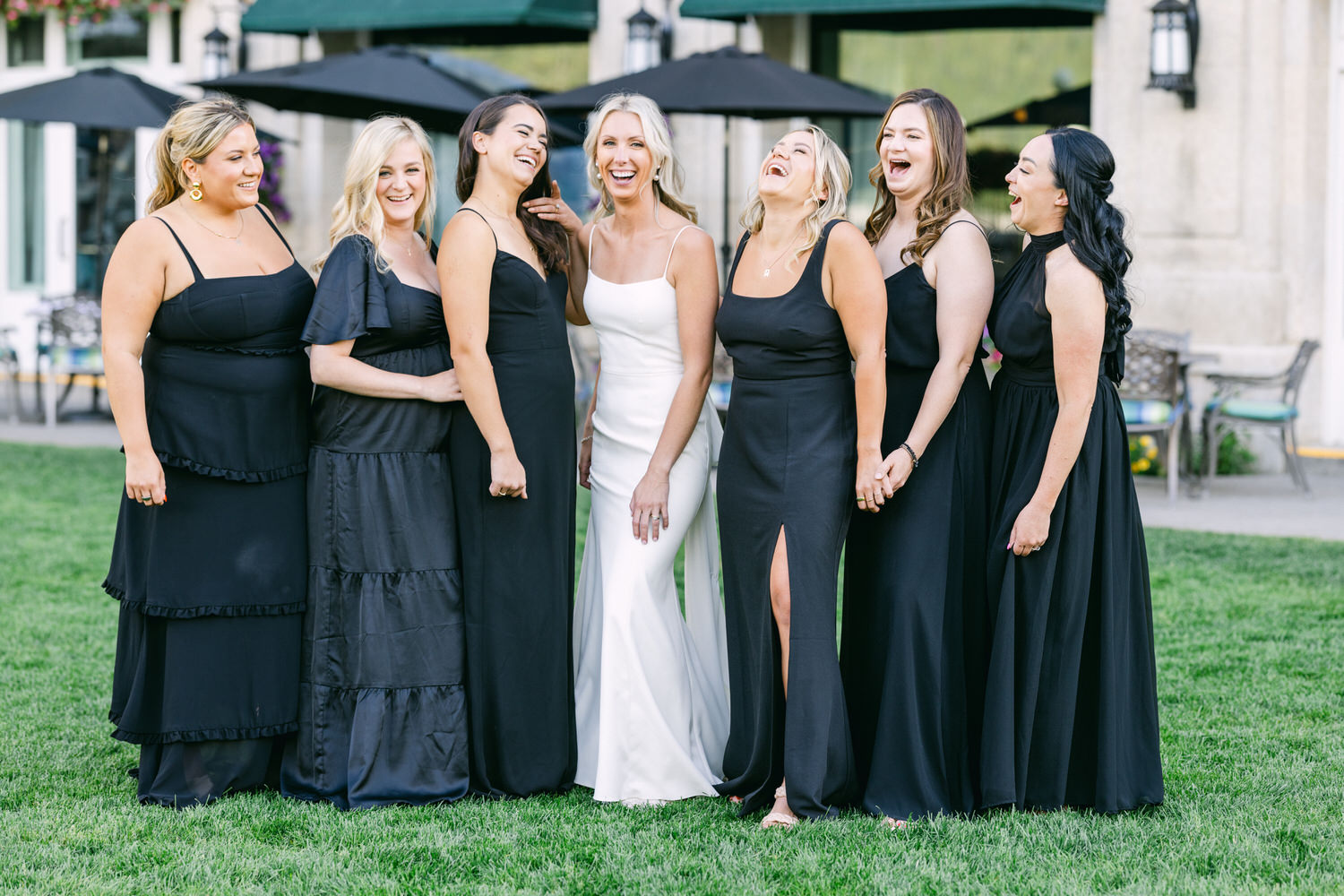 A bride in a white dress laughing with her bridesmaids in black dresses on a grassy lawn