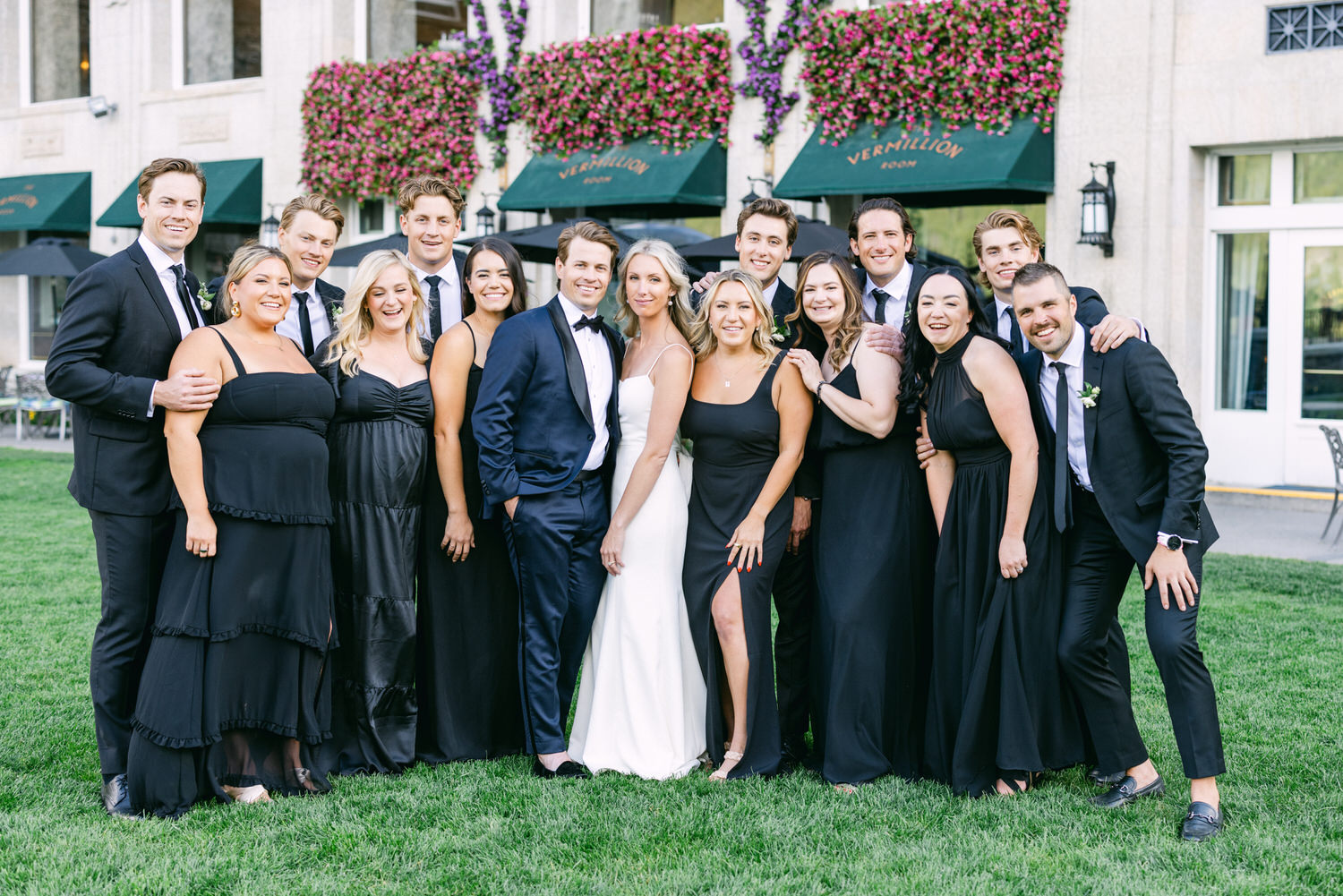 A group of people dressed in formal wear posing together on a lawn with a building adorned with flowering plants in the background.