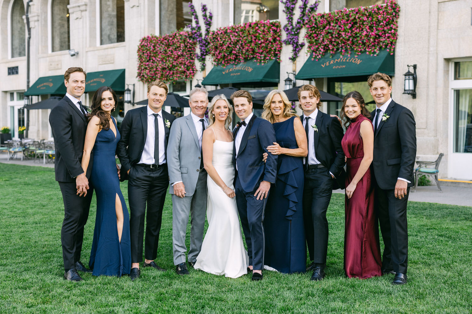 Group of well-dressed individuals posing for a wedding photo outside with a building adorned with flowers in the background