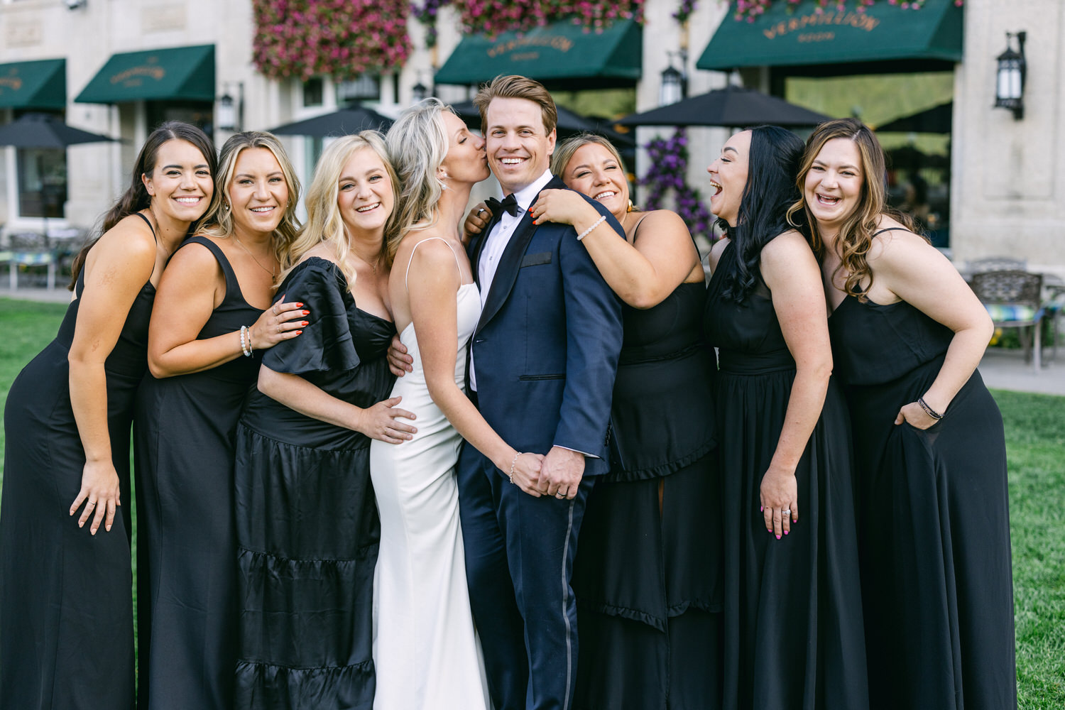 A group of smiling bridesmaids in black dresses embracing a happy bride and groom outdoors.