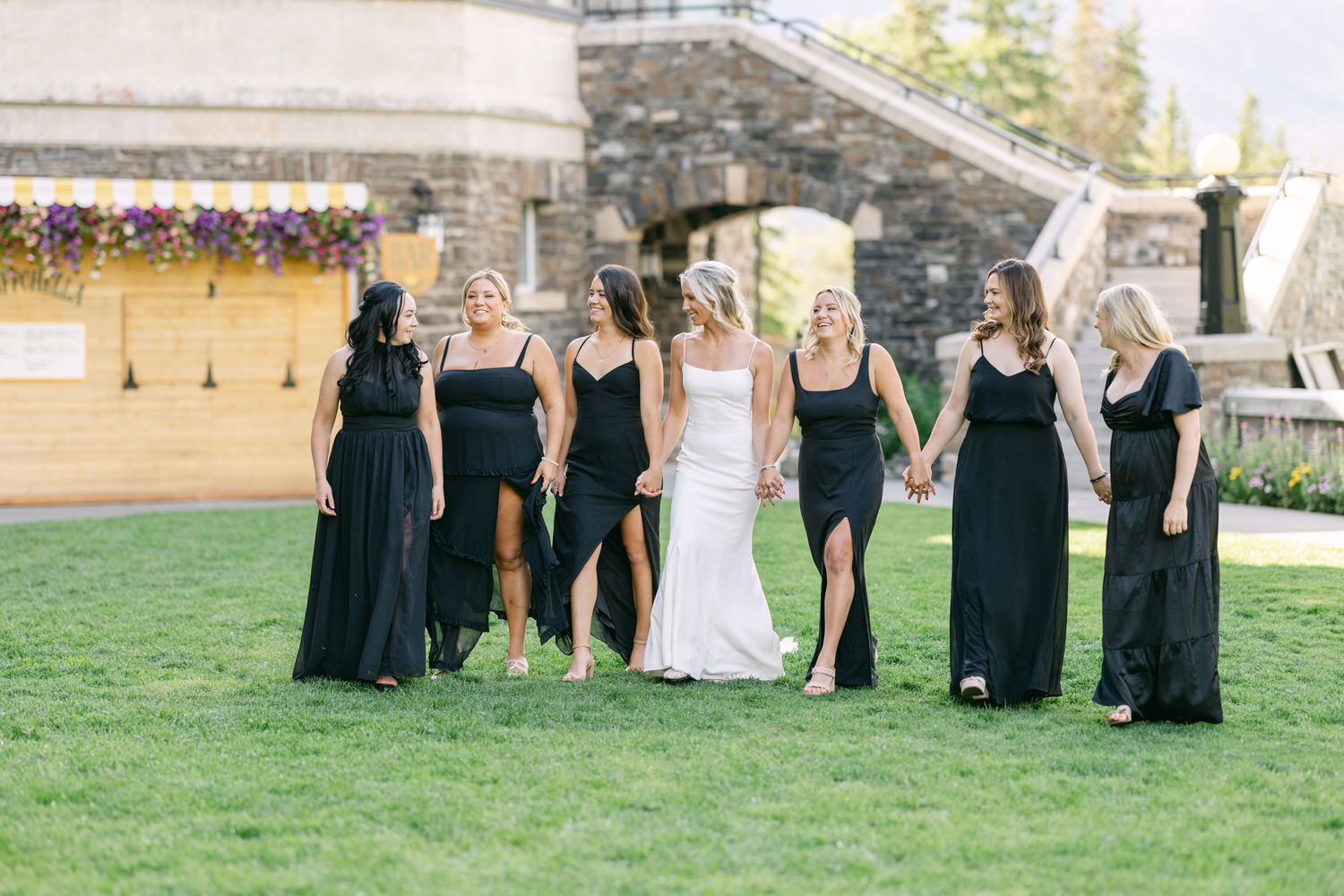 A bride in white and her bridesmaids in black dresses walking and laughing together on a grassy lawn.