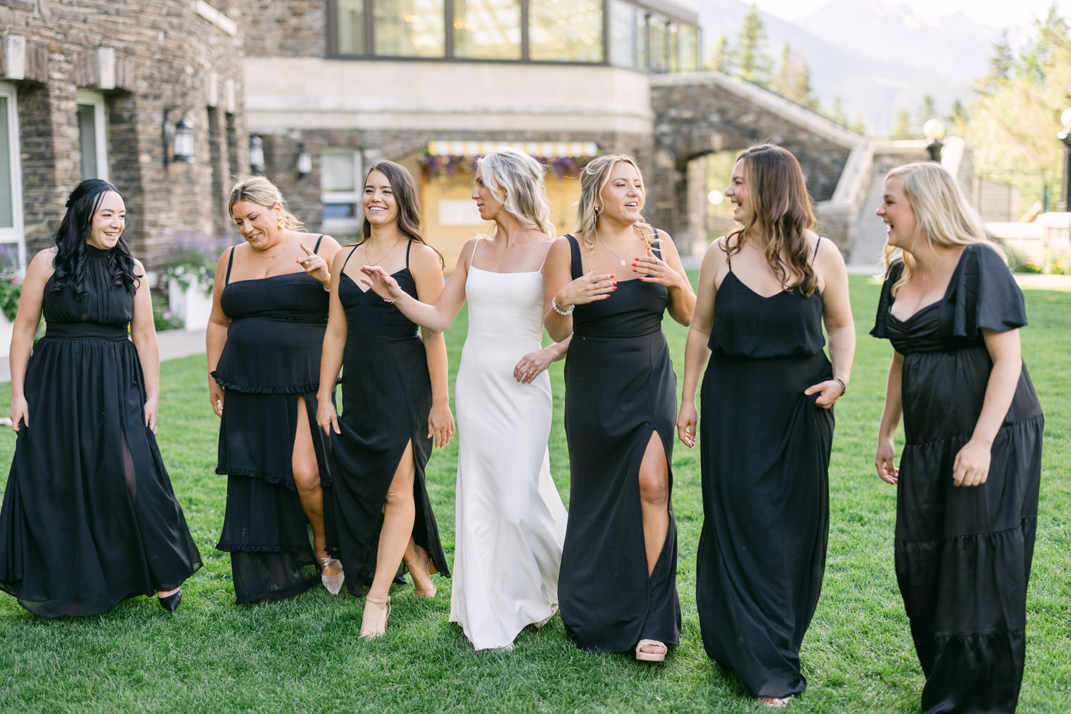 Group of women in black dresses with one in white, walking on grass and enjoying a moment together outside a stone building.