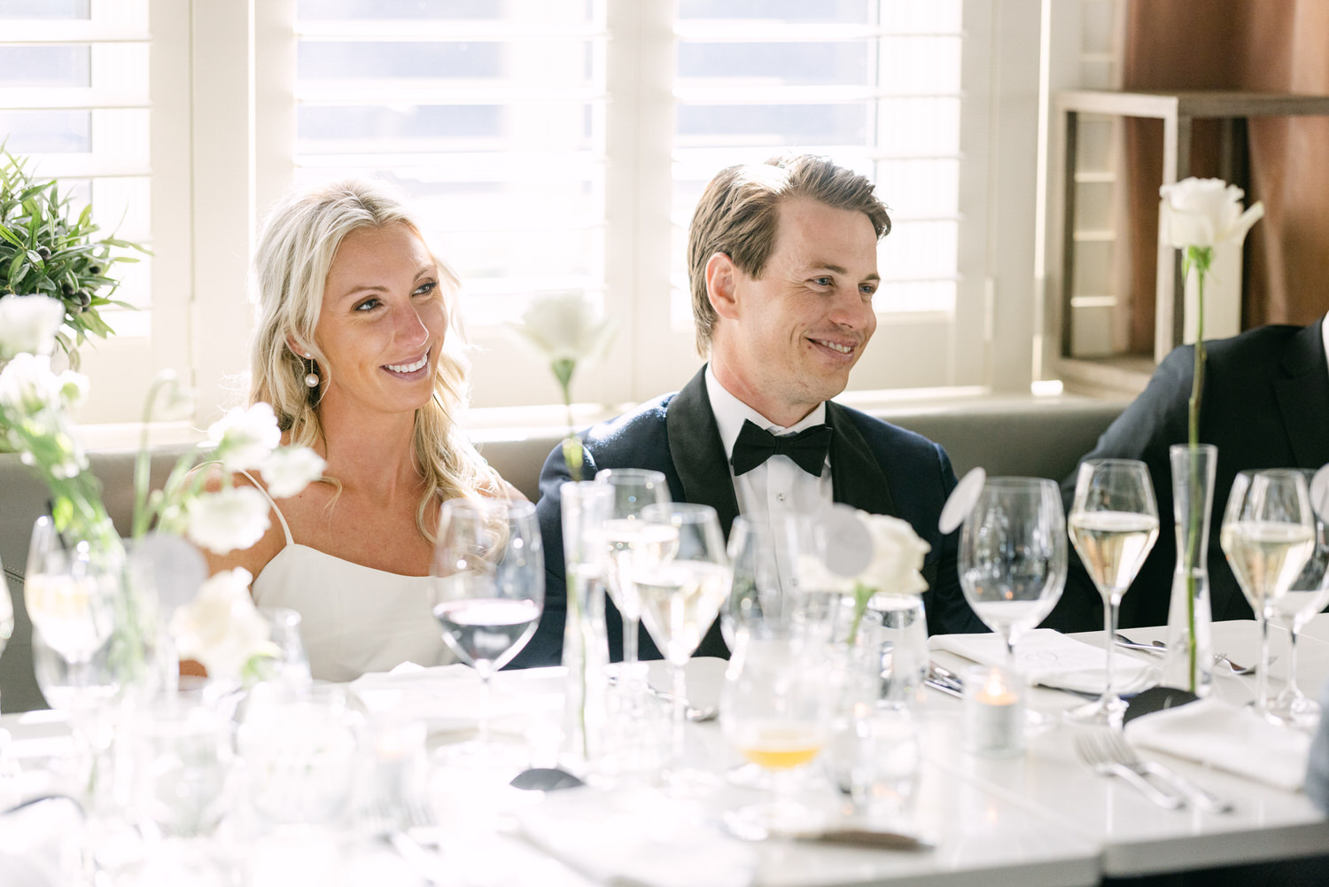  A smiling couple at a wedding reception table with white floral decorations and glasses of wine.