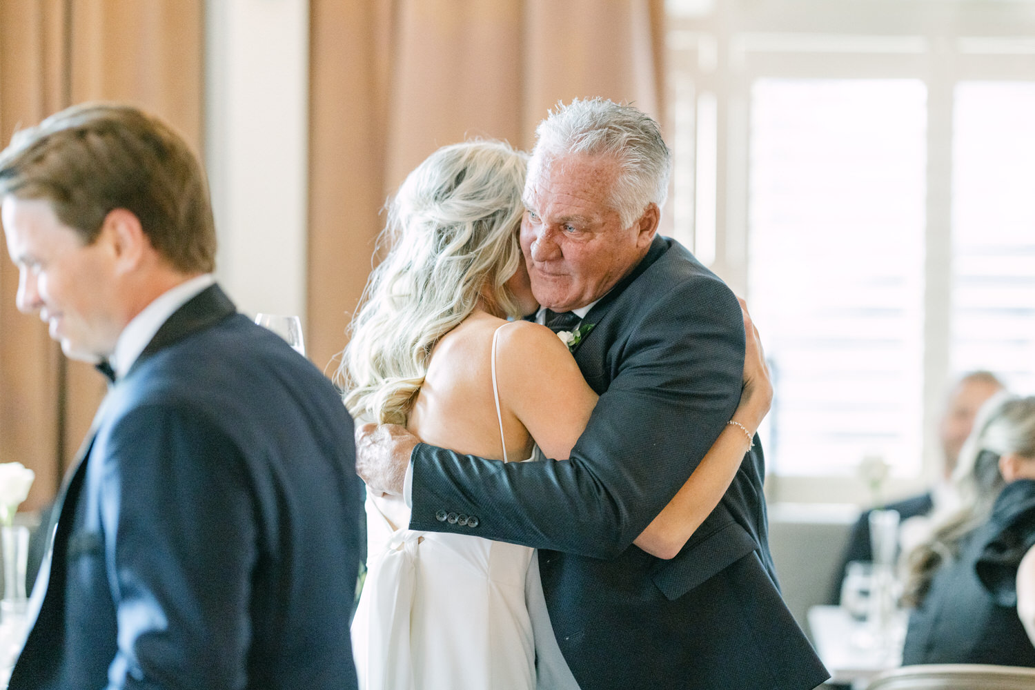 Elderly man sharing a heartfelt hug with a younger woman at a wedding reception, with other guests in the background.