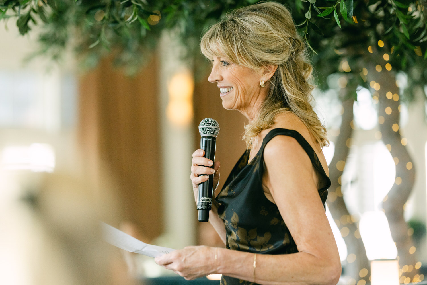 A smiling woman holding a microphone and a piece of paper while speaking at a decorated venue with bokeh lights in the background.