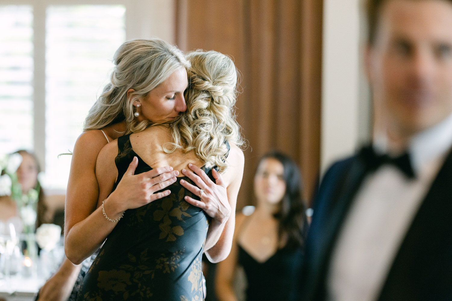 Two women hugging each other tightly at an event, with guests in the background and a man in the foreground out of focus.