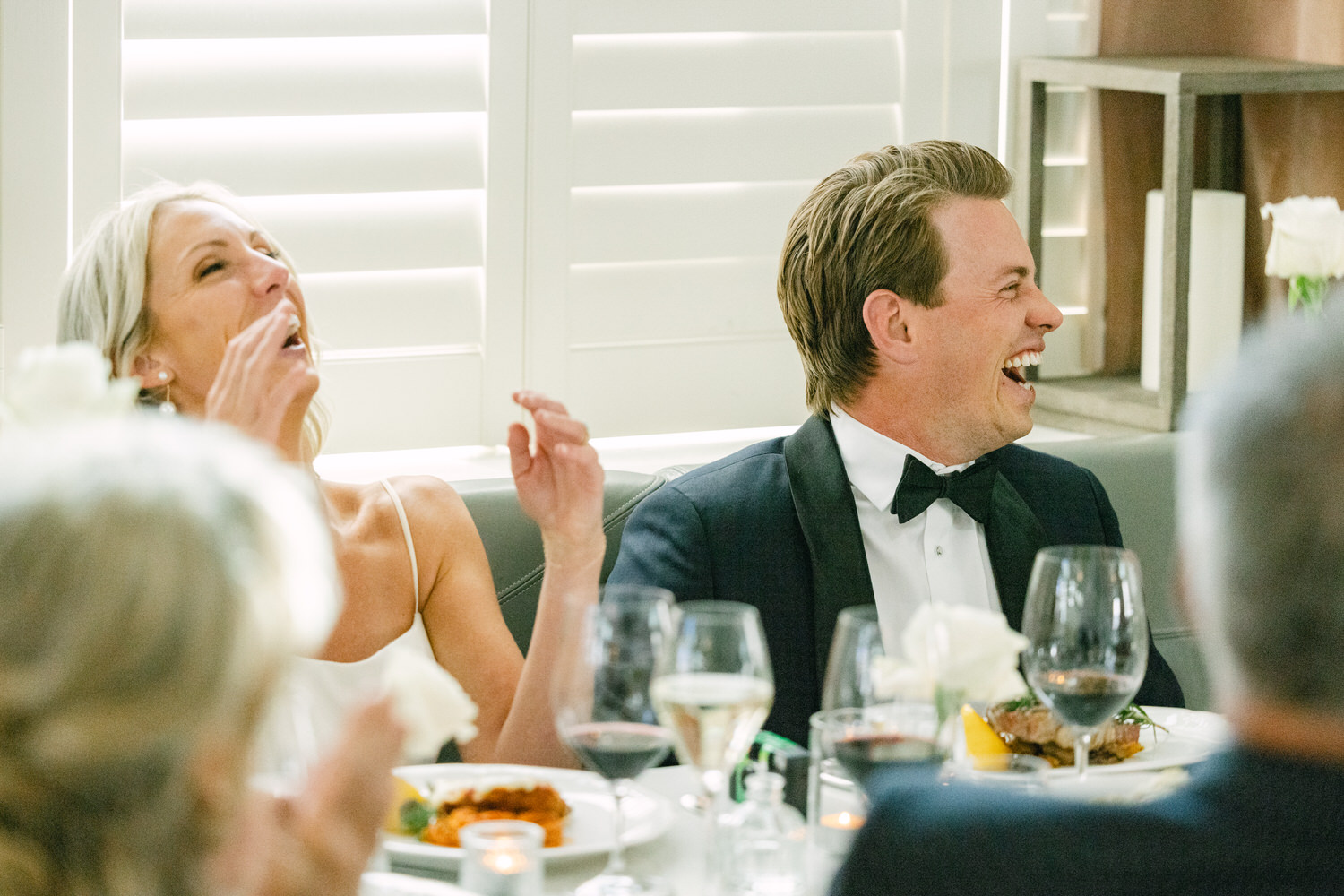 A woman and man in formal attire laughing joyfully at a banquet table with wine glasses and plates of food in the foreground.