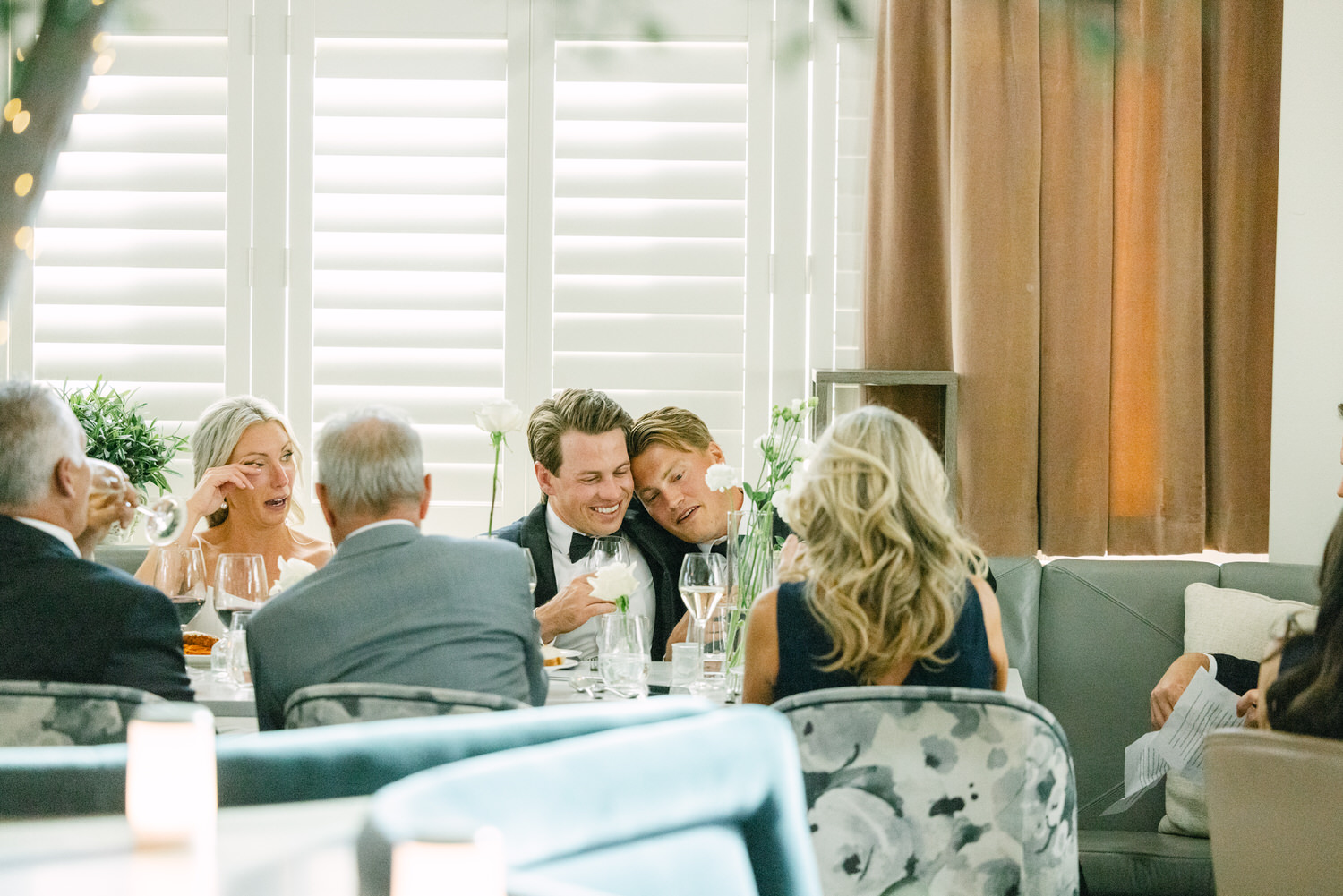 Group of people enjoying a conversation around a table with white roses in a well-lit, stylish venue.