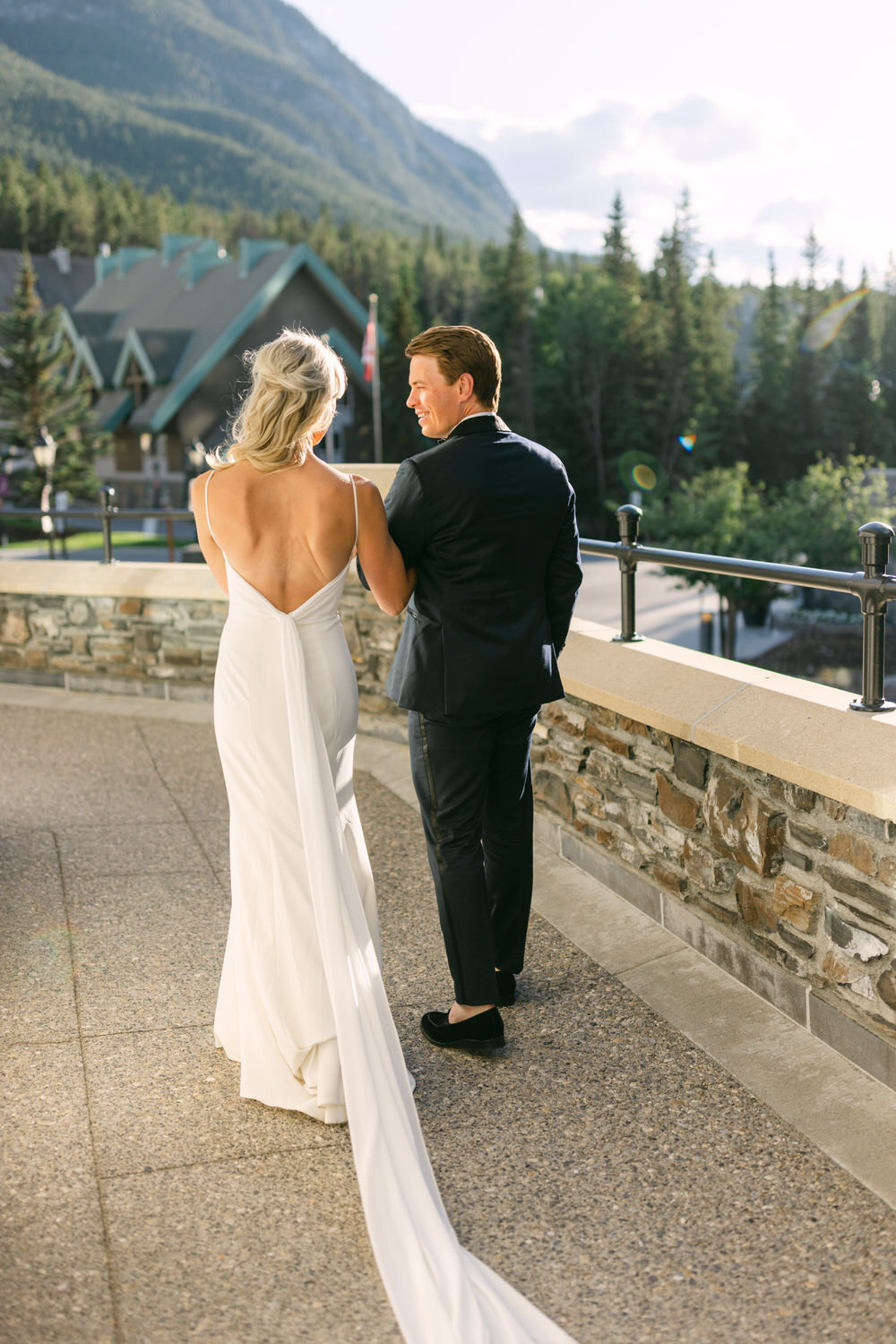 A couple in wedding attire standing on a terrace, with the bride in a white dress looking at the groom in a suit, with a scenic mountain backdrop.