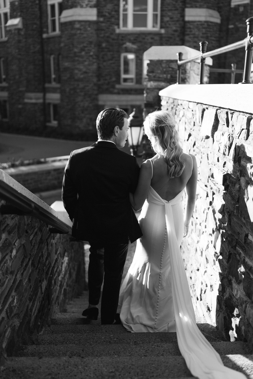 Black and white photo of a man in a suit and a woman in a bridal gown walking down stone stairs together