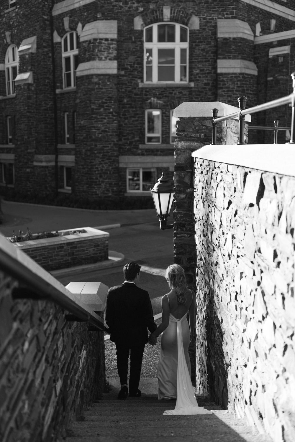 A black and white photograph of a couple holding hands while walking down stone steps, with a vintage street lamp and a brick building in the background.