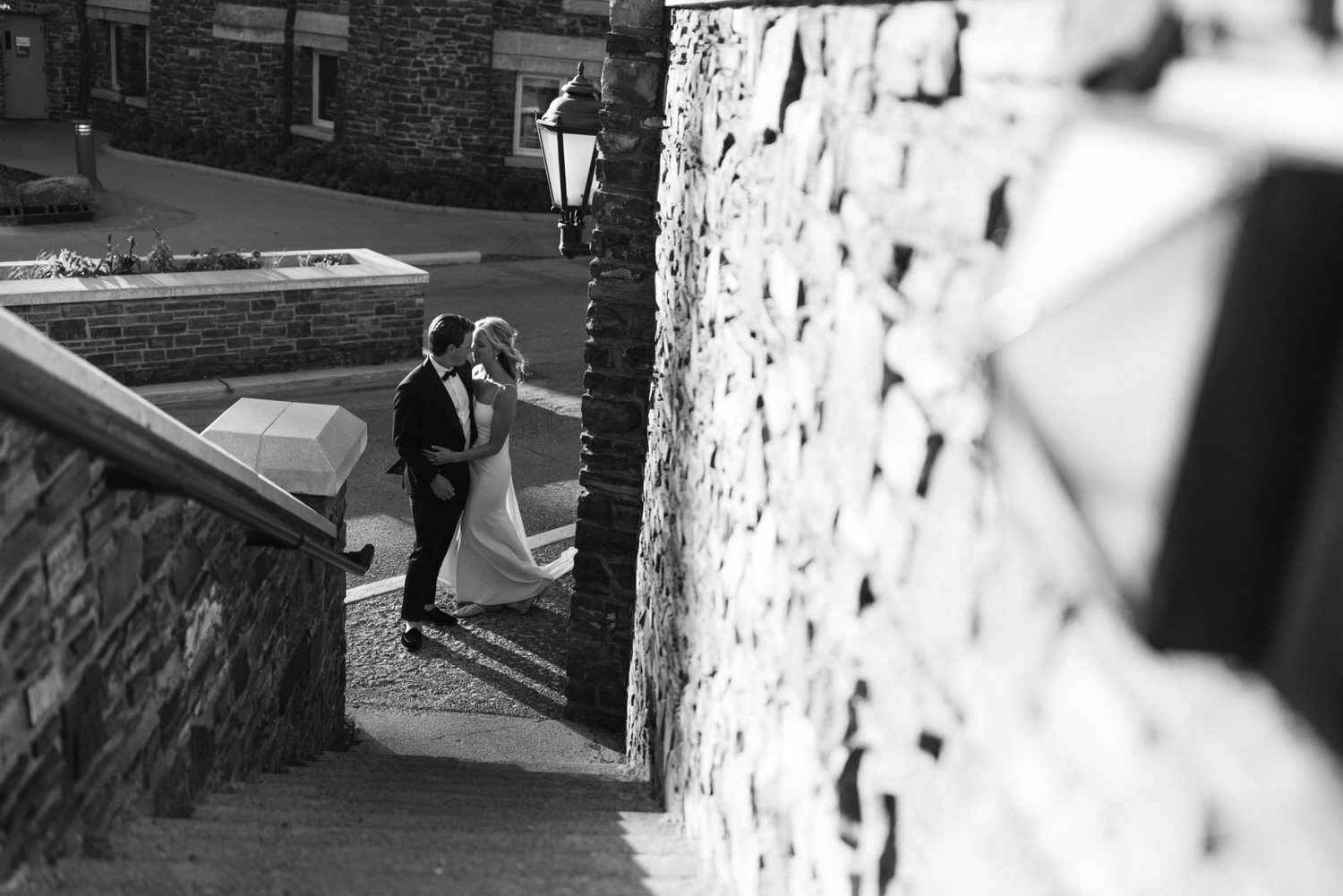 A black and white photo of a bride and groom sharing a kiss beside a stone wall, with a vintage street lamp and buildings in the background.