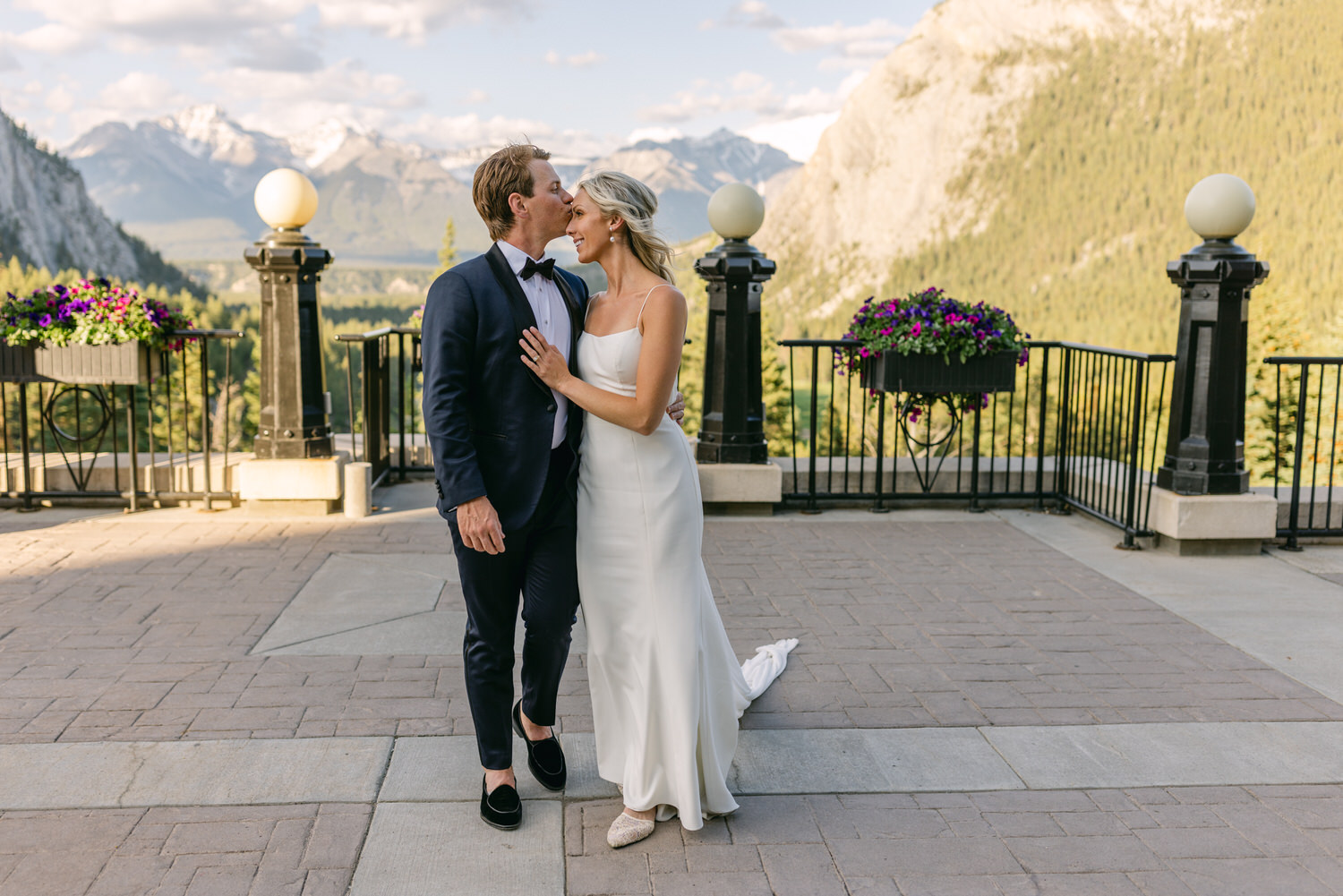 A bride and groom kissing on a balcony with a picturesque mountain background.