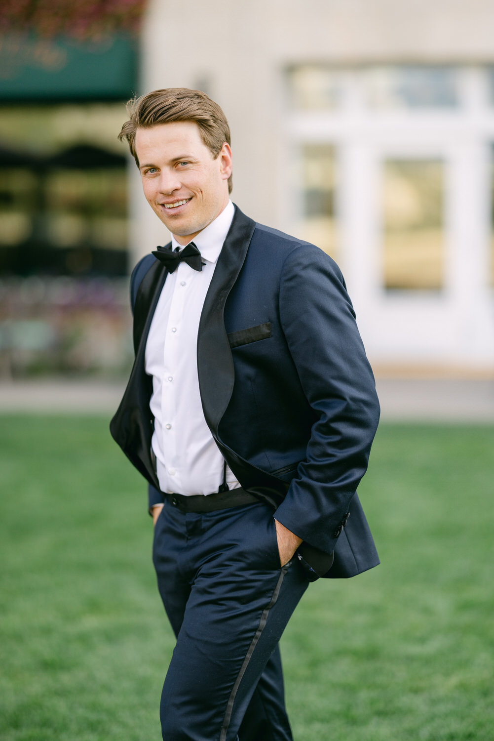 A man in a black tuxedo and bow tie smiling at the camera with a blurred background.