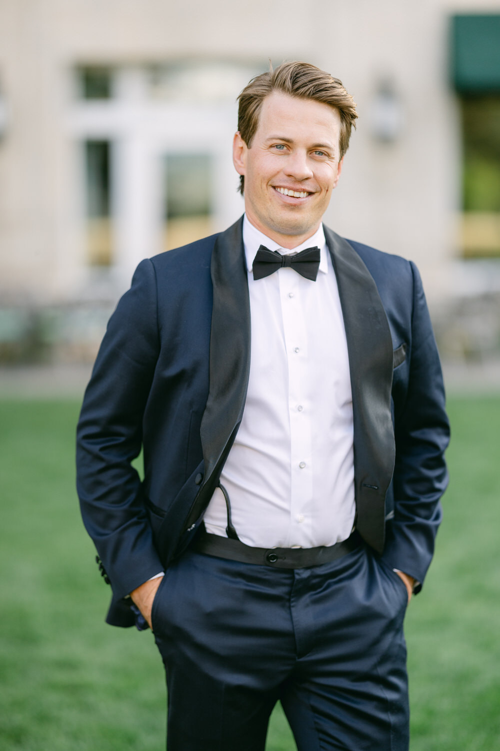 A man dressed in a black tuxedo, white dress shirt, and bow tie, smiling with his hands in pockets, standing outdoors.