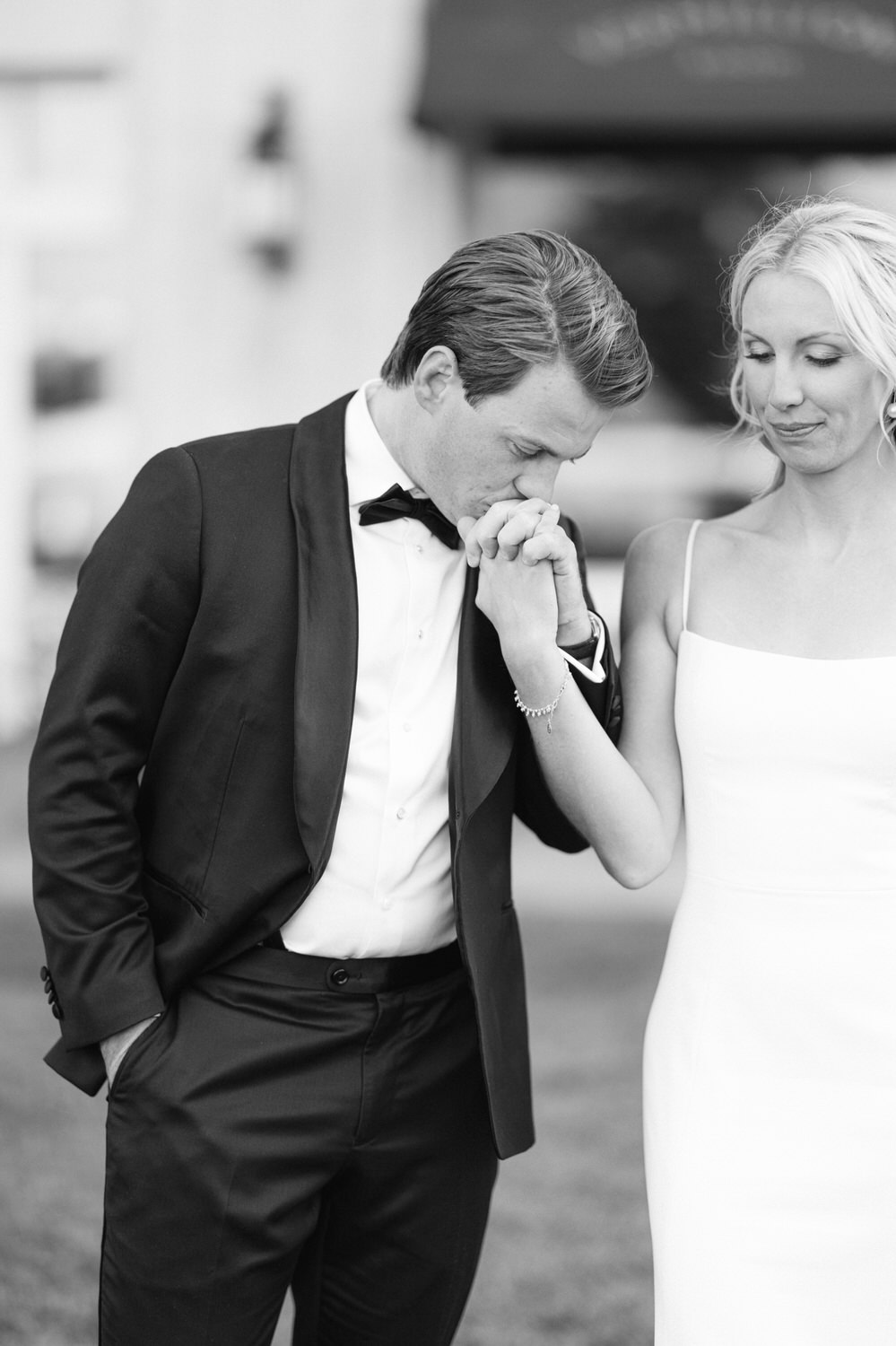 Black and white image of a groom kissing the bride's hand, both dressed in wedding attire, expressing a moment of affection.