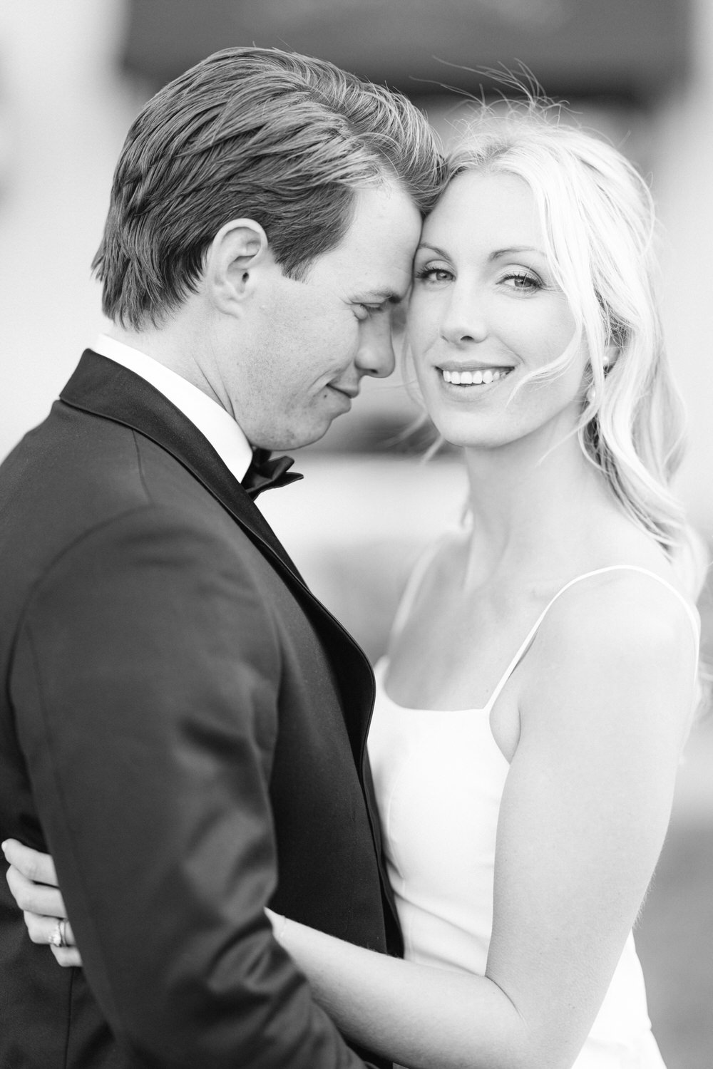 Intimate Couple Embrace:::A black and white photo of a man in a tuxedo with his forehead against a smiling woman in a white dress.