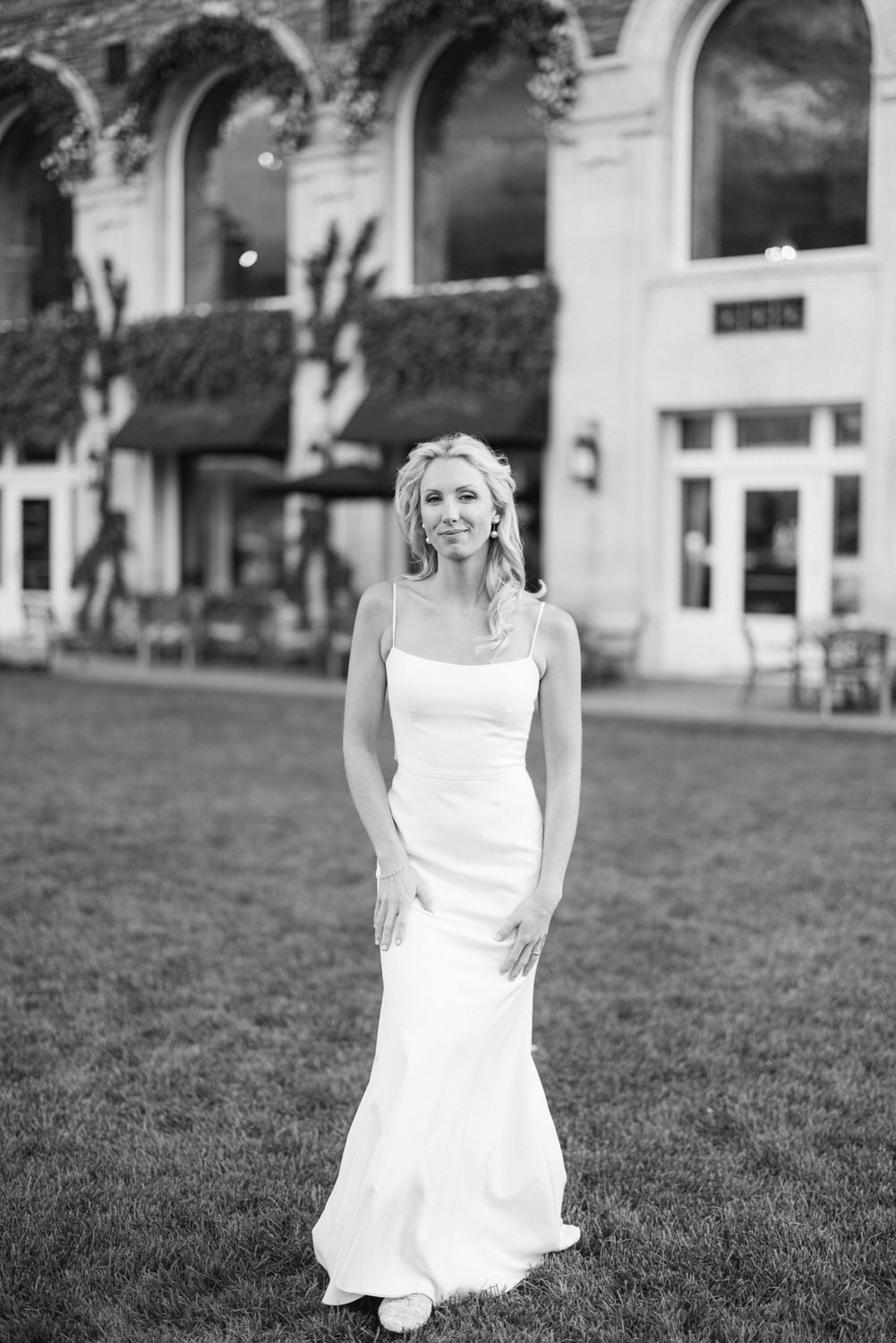 A woman in a white dress standing in front of a building with arched windows, captured in black and white.