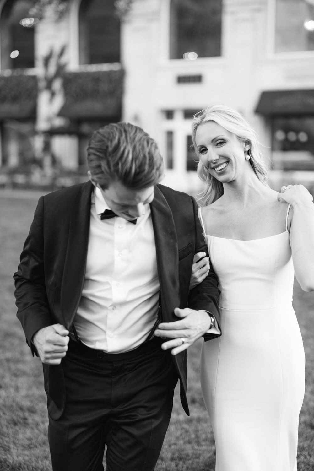 Black and white photo of a smiling woman in an elegant dress beside a man adjusting his suit, both in formal attire, with a blurred background suggesting an upscale outdoor setting.