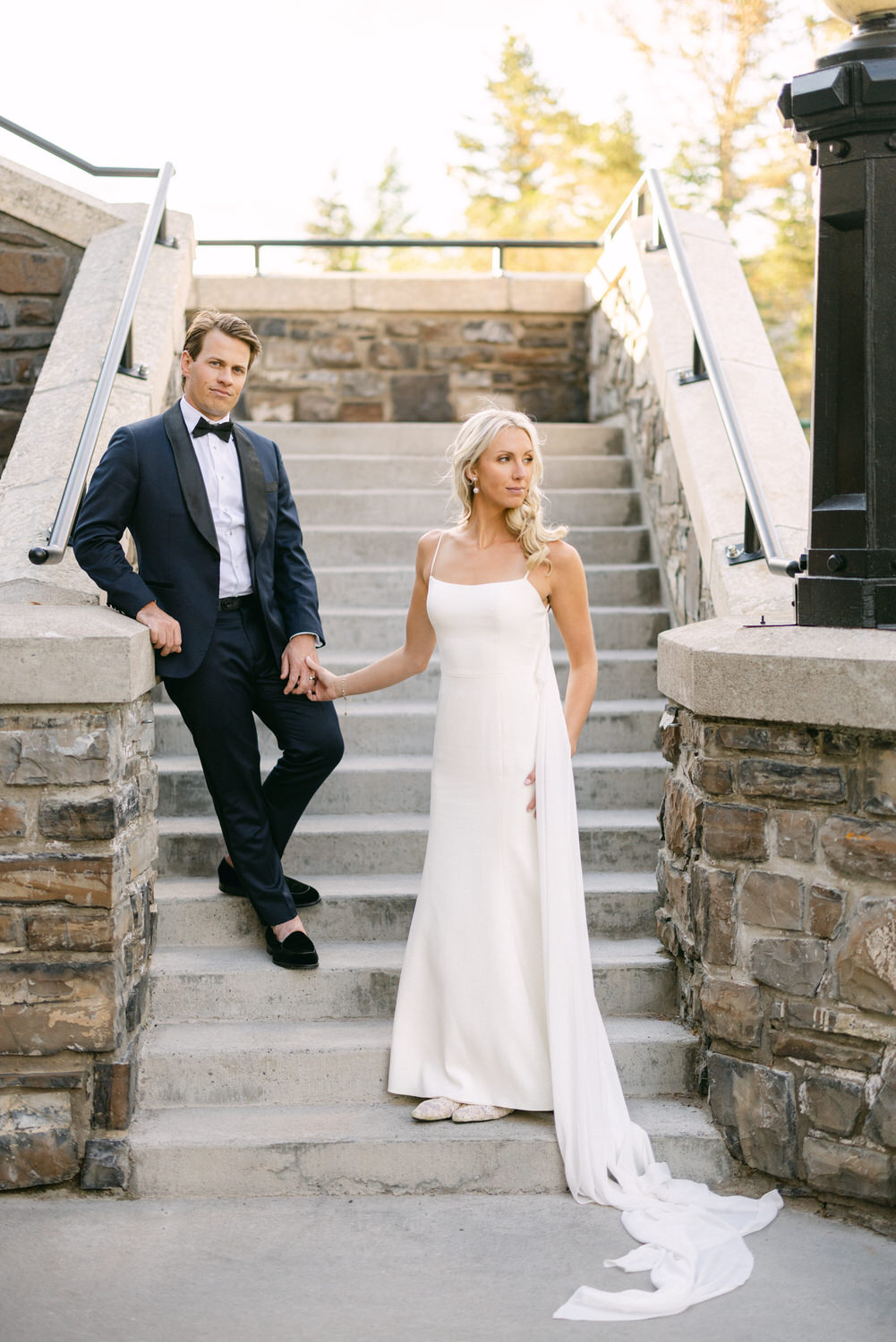 A man in a suit and a woman in a wedding dress holding hands on a stone staircase, with a tranquil outdoor background.