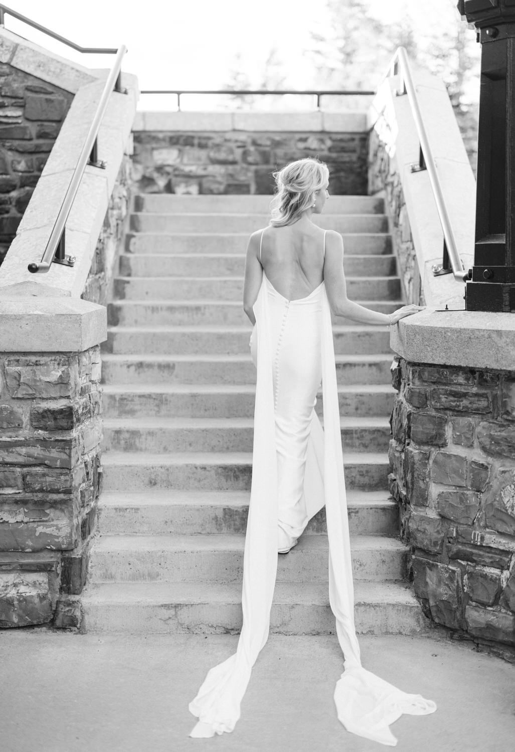 A black and white image of a woman in an elegant dress climbing stairs, viewed from behind.