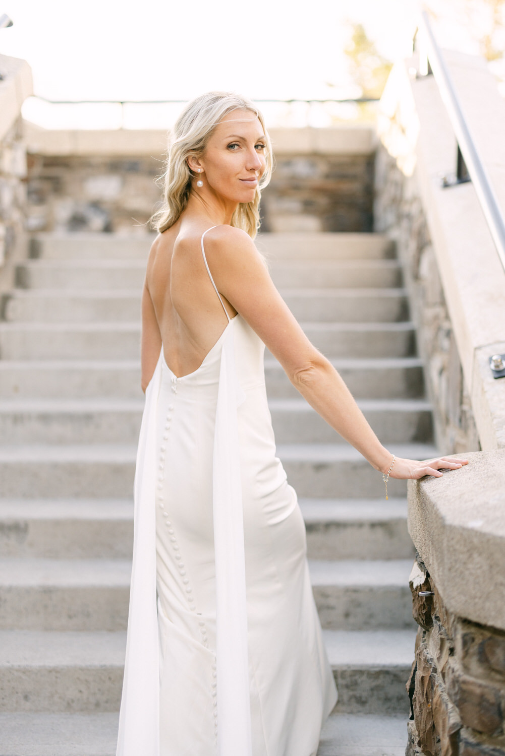 A woman in a chic white wedding dress looking over her shoulder while standing on stone stairs.