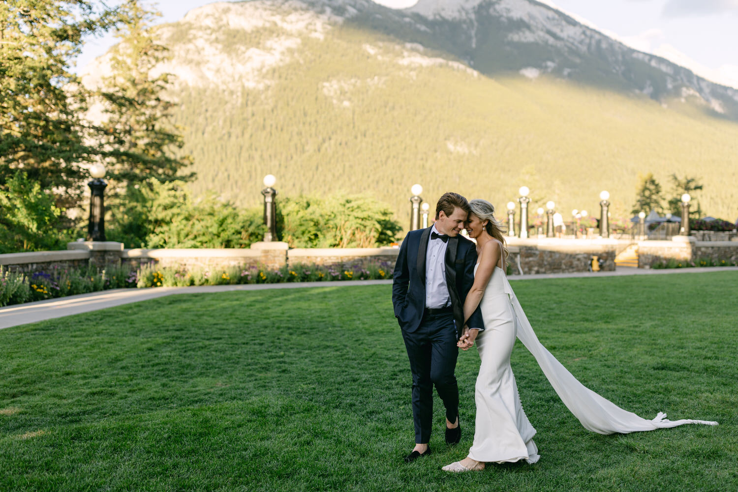 A couple in formal wedding attire sharing an intimate moment in a lush garden with mountainous backdrop.