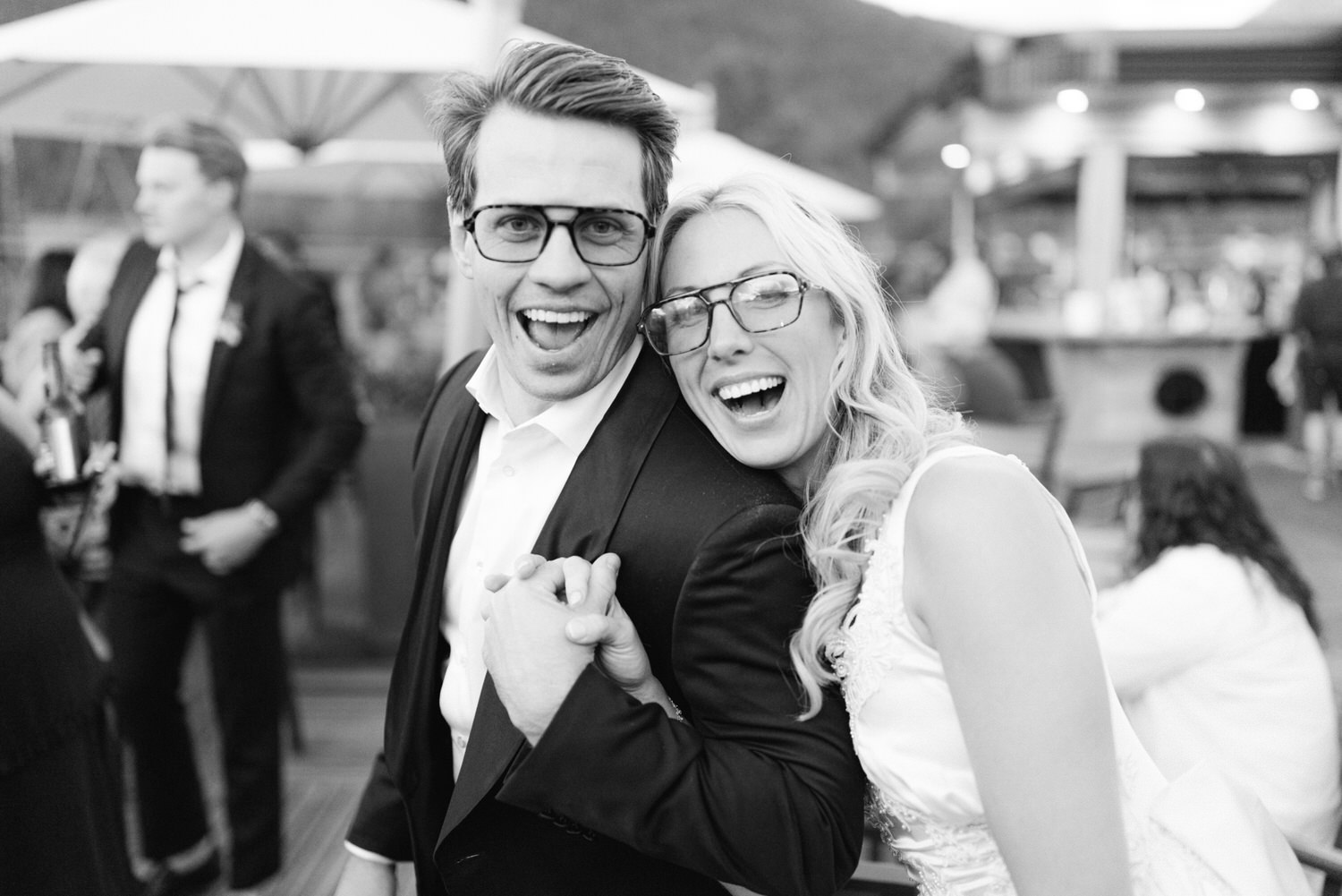 A man and woman laughing together joyfully, both wearing glasses and formal attire, in a blurry black and white party setting.