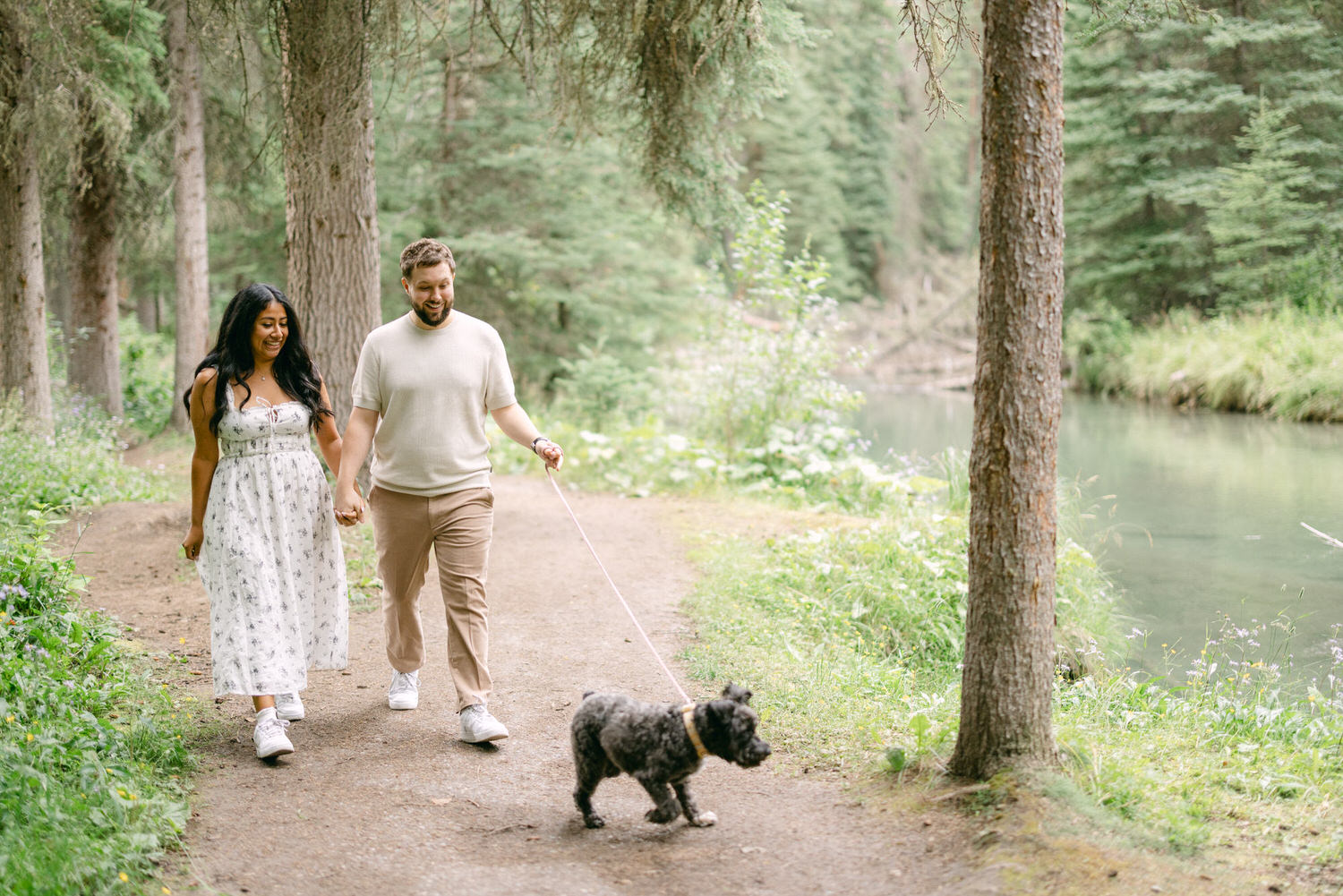 A man and woman holding hands and smiling while walking a small dog on a leash along a forest trail next to a river.