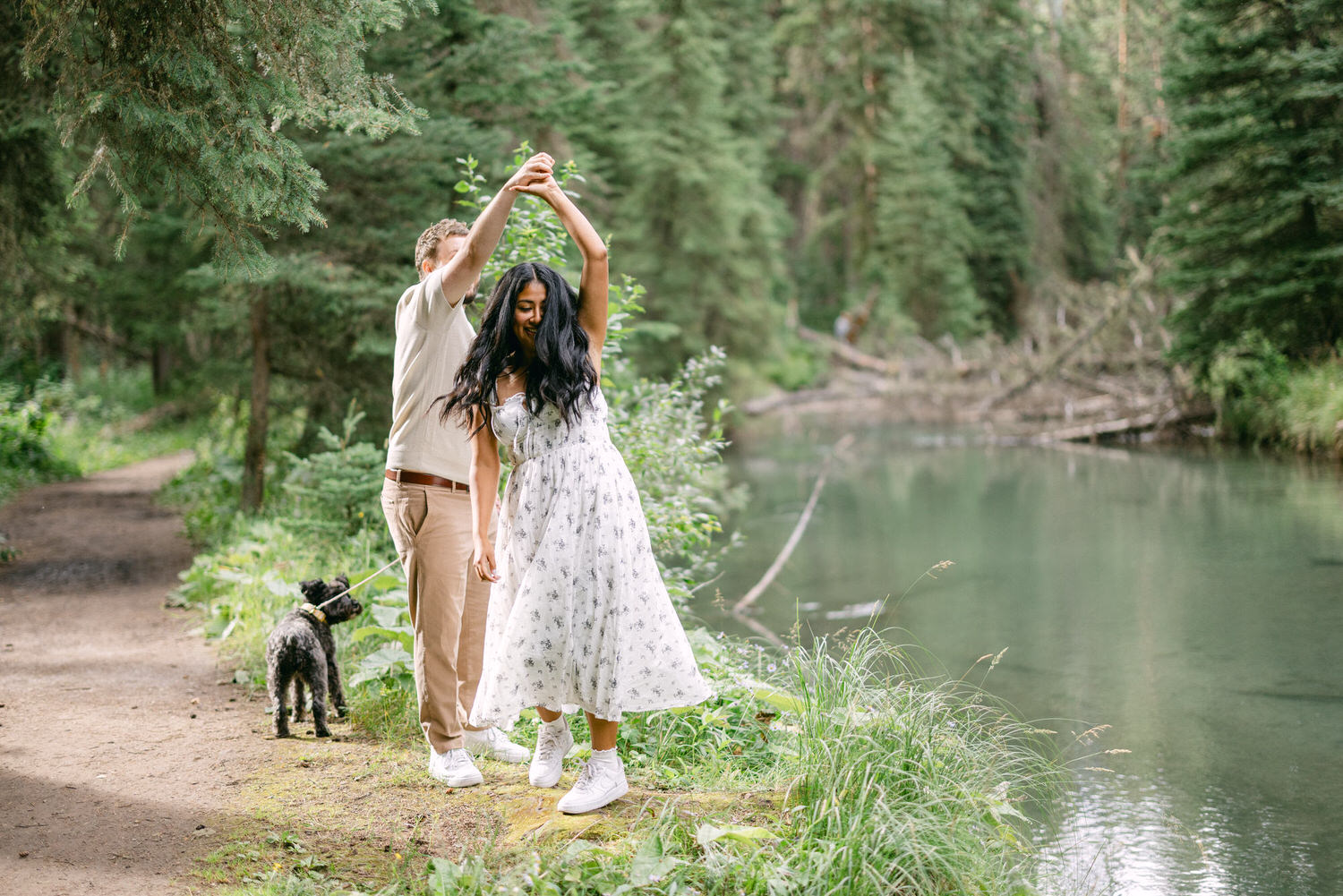 A couple playfully dancing by a serene lakeside, with a dog on a leash nearby in a lush forest setting.