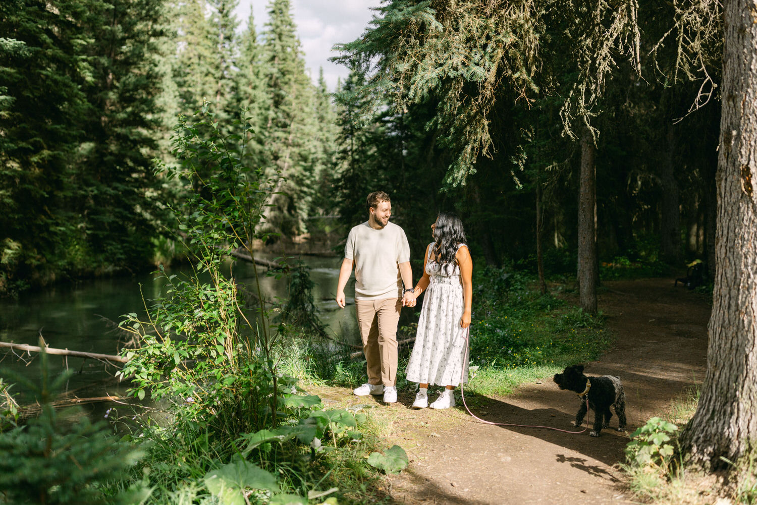 A couple holding hands on a tranquil walk through a lush forest trail with their black dog on a leash standing beside them.