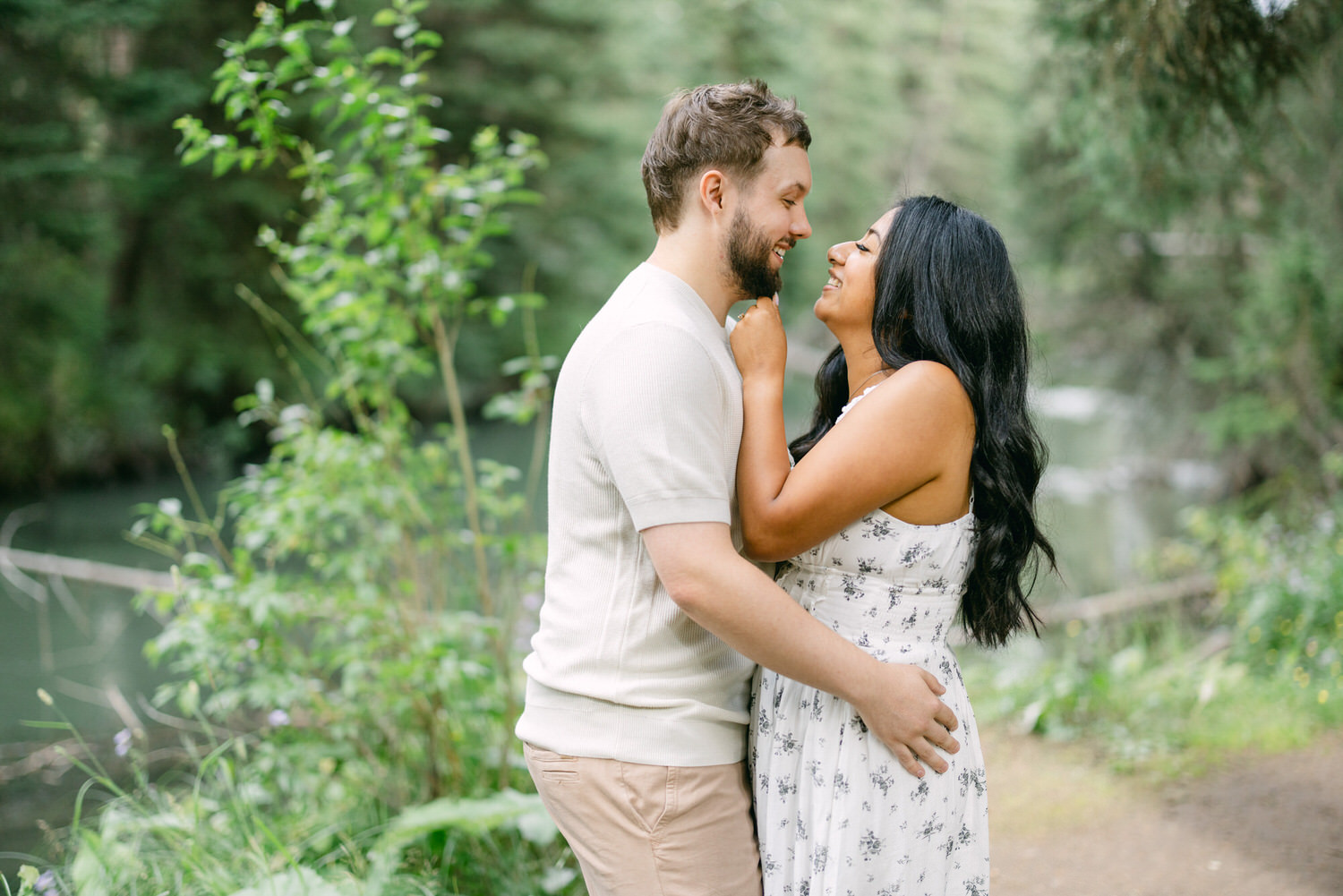 A man and woman embracing and looking at each other affectionately in a serene outdoor setting with greenery and a water body in the background.