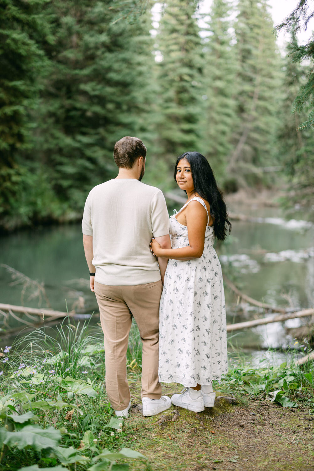 A couple standing together in a forest, with the woman looking back over her shoulder at the camera while the man’s back is turned, near a serene river.