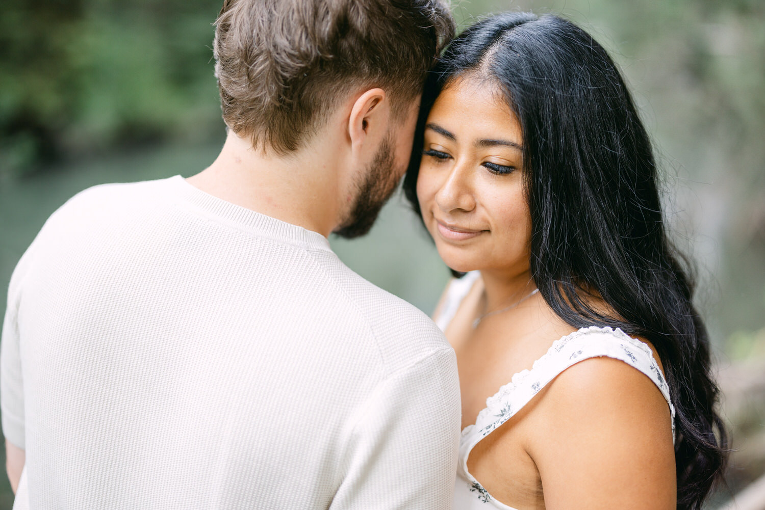 A woman with a serene expression leaning her head close to a man, both standing in a natural setting with selective focus.