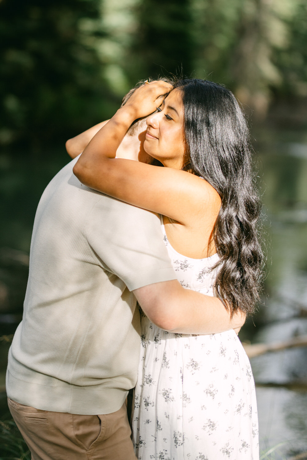 A woman in a floral dress with long hair embracing a person outdoors near a river, surrounded by natural greenery.