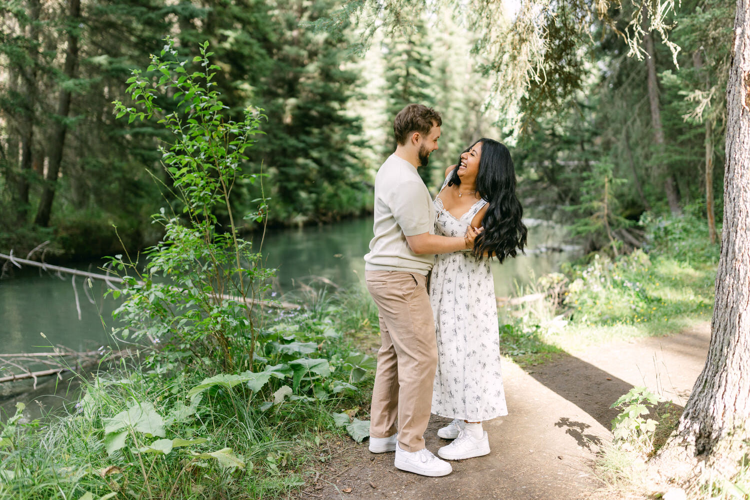 A man and woman standing close and looking at each other affectionately beside a serene forest stream, surrounded by lush greenery.