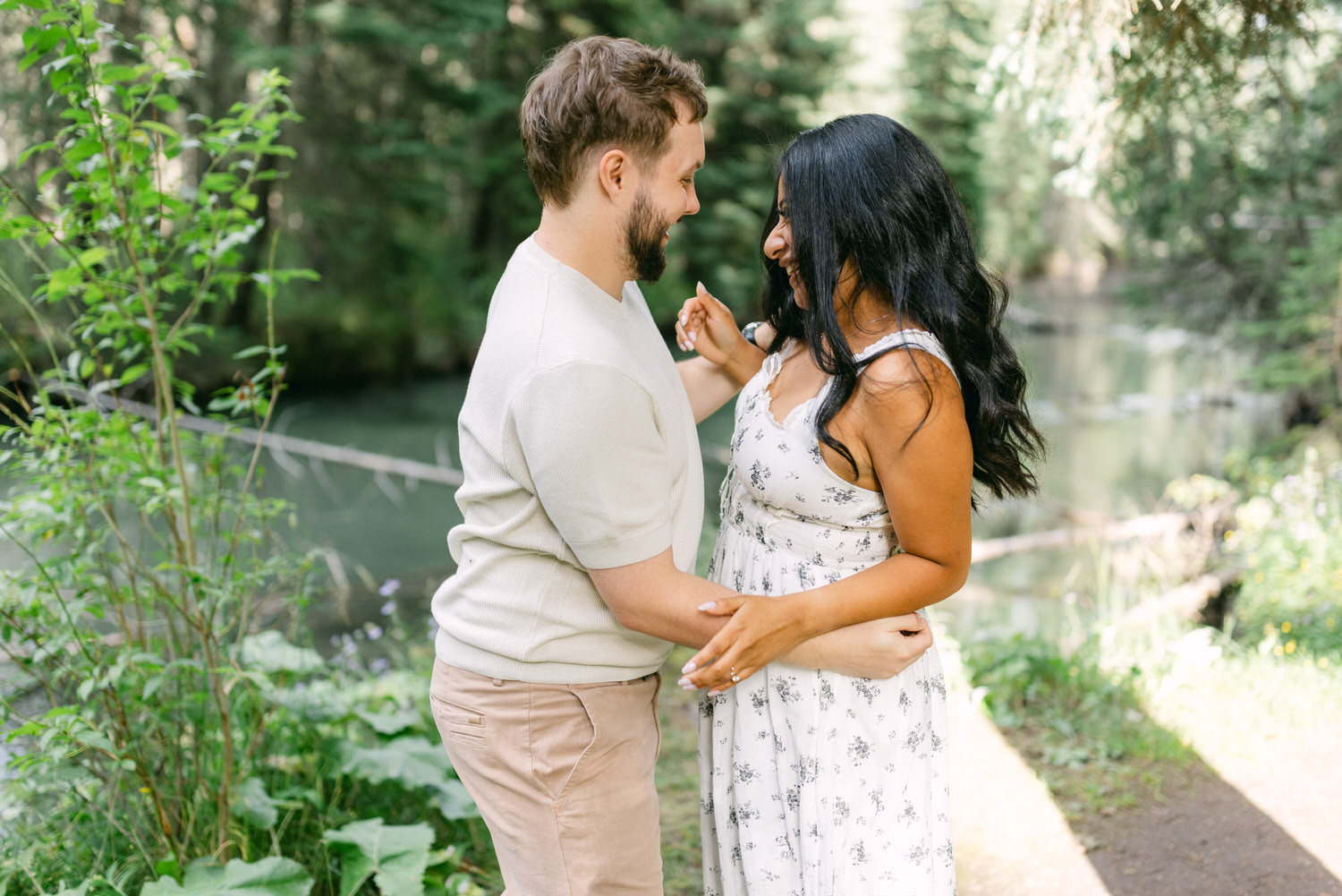 A man and woman smiling at each other while holding hands outdoors, with a serene river and lush greenery in the background.